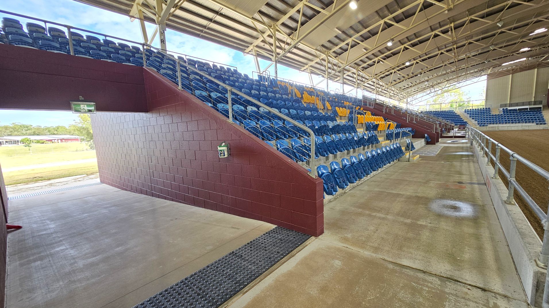 A covered stadium seating area with blue and yellow plastic seats arranged in rows. On the left, a maroon brick wall supports the seating structure, with an exit sign above a passageway. The right side features a concrete walkway with metal railings, leading to an open area with a dirt floor. The ceiling is made of metal beams and panels, providing shelter. In the background, there is a grassy area and a building visible through the open sides.