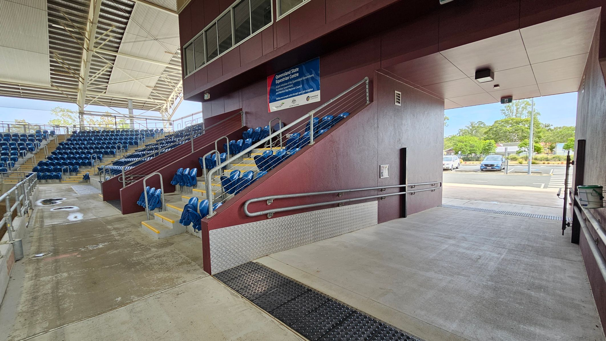 On the left, there are rows of blue stadium seats arranged on tiered steps, with metal railings separating sections. The ceiling is covered with a metal structure. In the centre, a maroon wall with a sign and a ramp with handrails leads downwards. On the right, an open passageway leads outside, where a few cars are parked and trees are visible. The floor is a mix of concrete and textured surfaces.