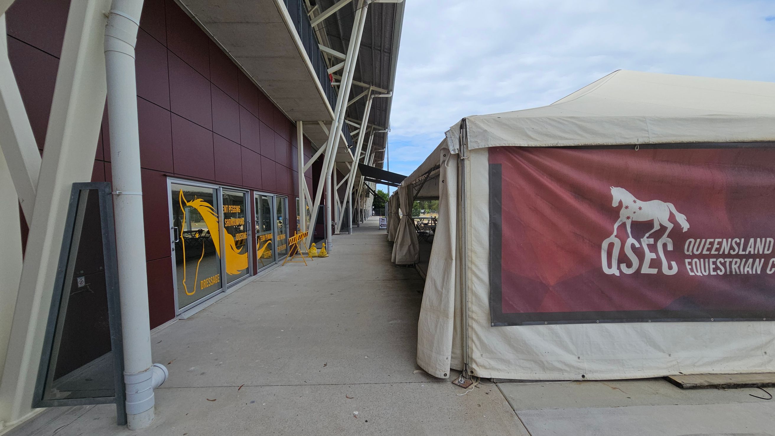 A walkway with a maroon building on the left featuring large windows with yellow horse graphics and text. The building has a grey roof supported by white beams. On the right, a white tent with a maroon banner displaying "QSEC Queensland Equestrian Centre" and a horse graphic. The ground is concrete, and the area is open with some barriers and signs visible in the distance.