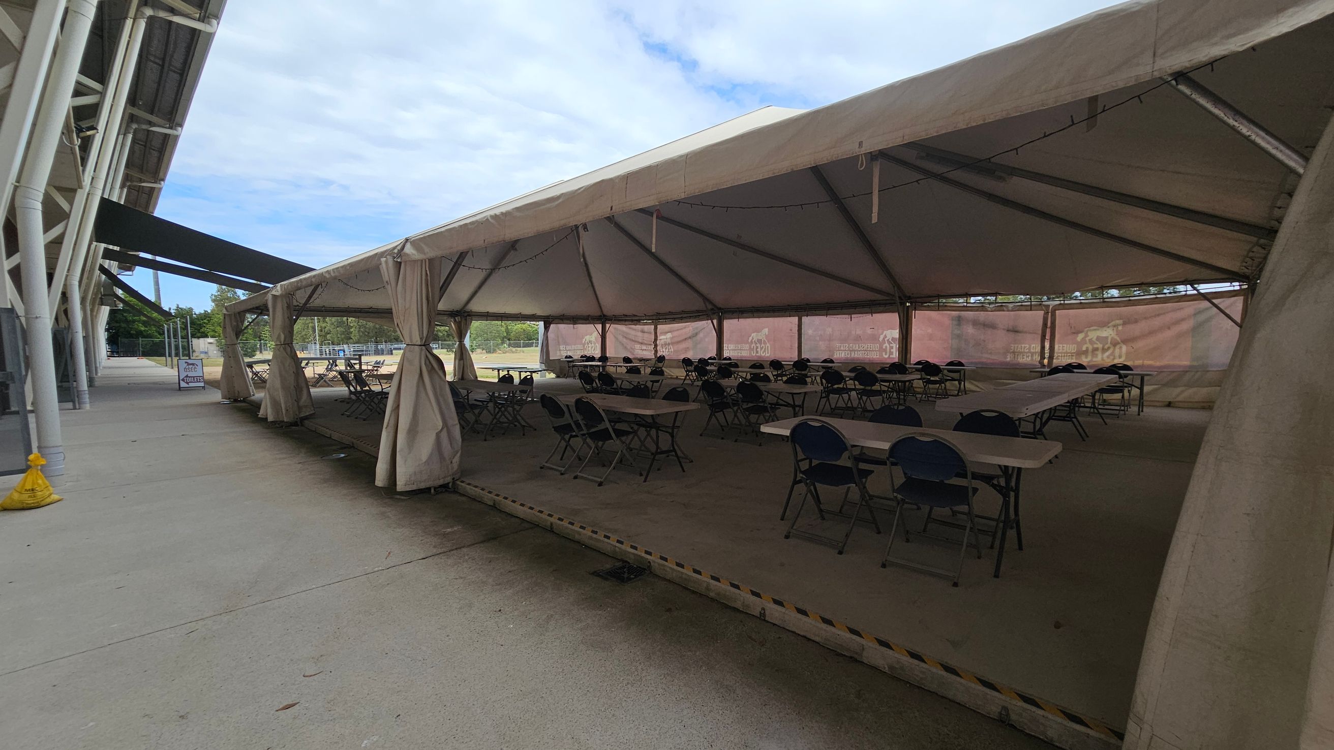A large outdoor tent with a beige canopy is set up on a concrete surface. Inside, there are several rows of folding tables and chairs arranged neatly. The tent is supported by metal poles and has open sides with curtains tied back. To the left, a building with a metal structure and a walkway is visible. The ground is a mix of concrete and green artificial turf. In the background, there are trees and a partially visible sign.