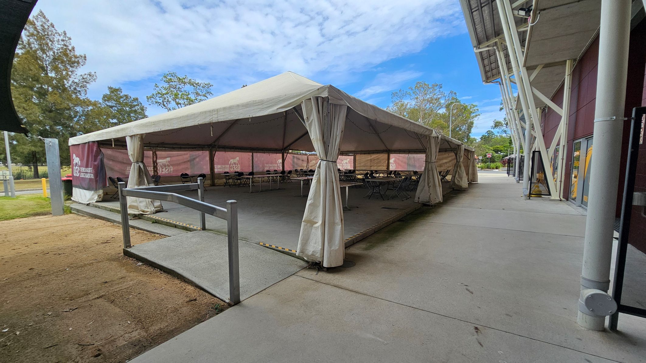 A large white tent is set up on a concrete surface, with open sides revealing tables and chairs inside. The tent is supported by metal poles and has a ramp leading up to it. To the right, a modern building with a red exterior and large windows is visible, featuring structural beams. The ground is a mix of concrete and grass, with trees in the background. The sky is clear and blue.