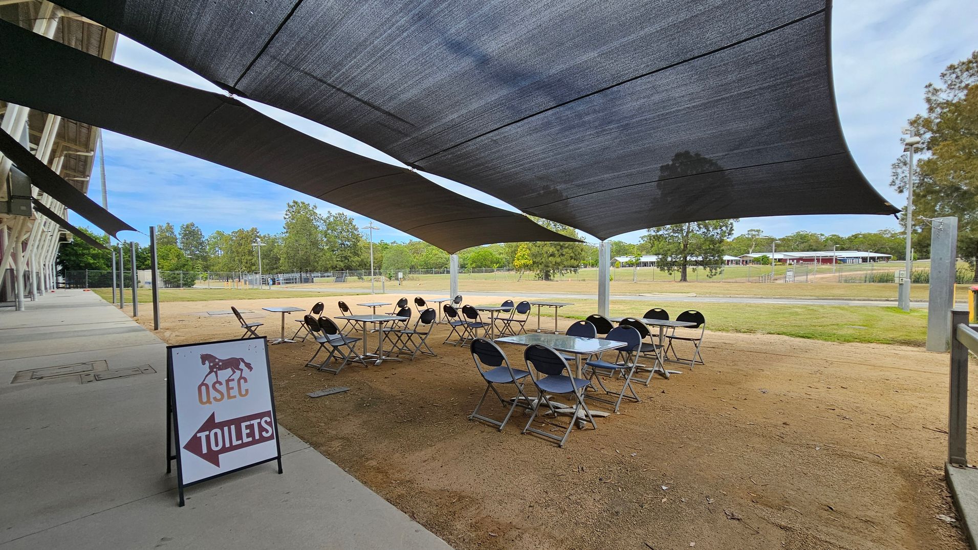 On the left, a sign with "QSEC TOILETS" and an arrow points left. Several tables and chairs are arranged on a sandy surface under large, dark shade sails. In the background, a grassy area with scattered trees and a distant building is visible. The foreground shows a concrete walkway alongside the seating area.