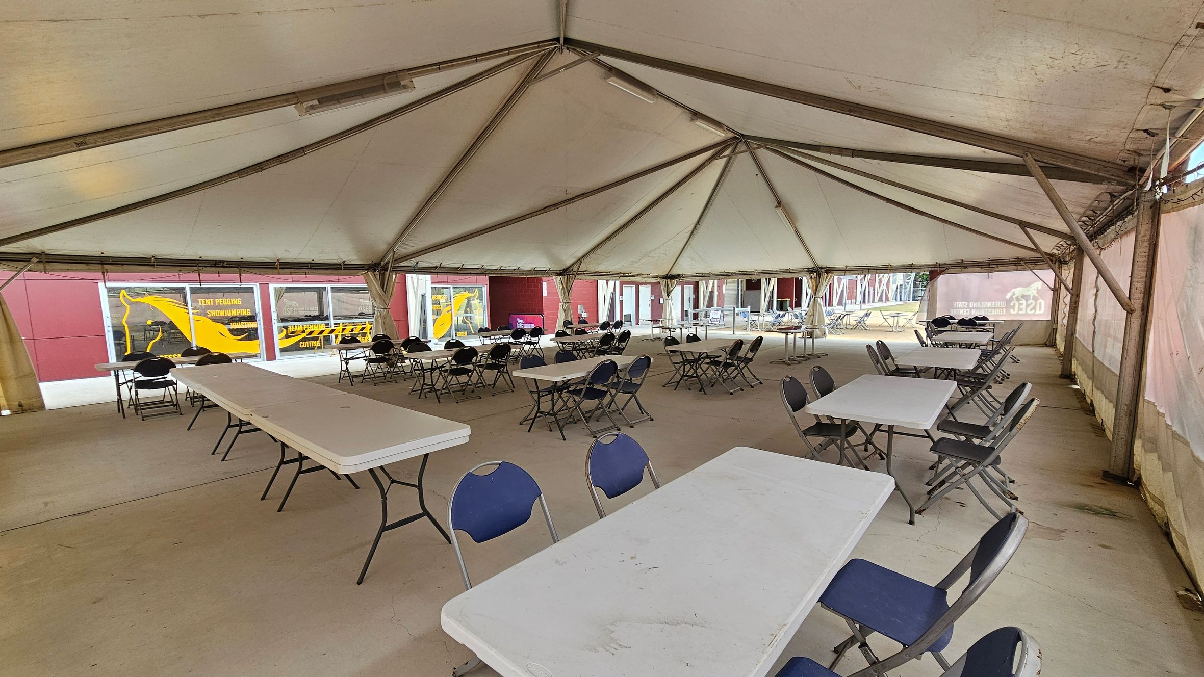 Inside a large tent with a high, peaked ceiling supported by metal beams. The floor is concrete. Several rectangular tables are arranged in rows, each surrounded by folding chairs with blue seats. The tent walls are partially transparent, revealing a red building outside with yellow graphics, including a horse silhouette and text. The space appears set up for an event or gathering, with ample seating and open areas.