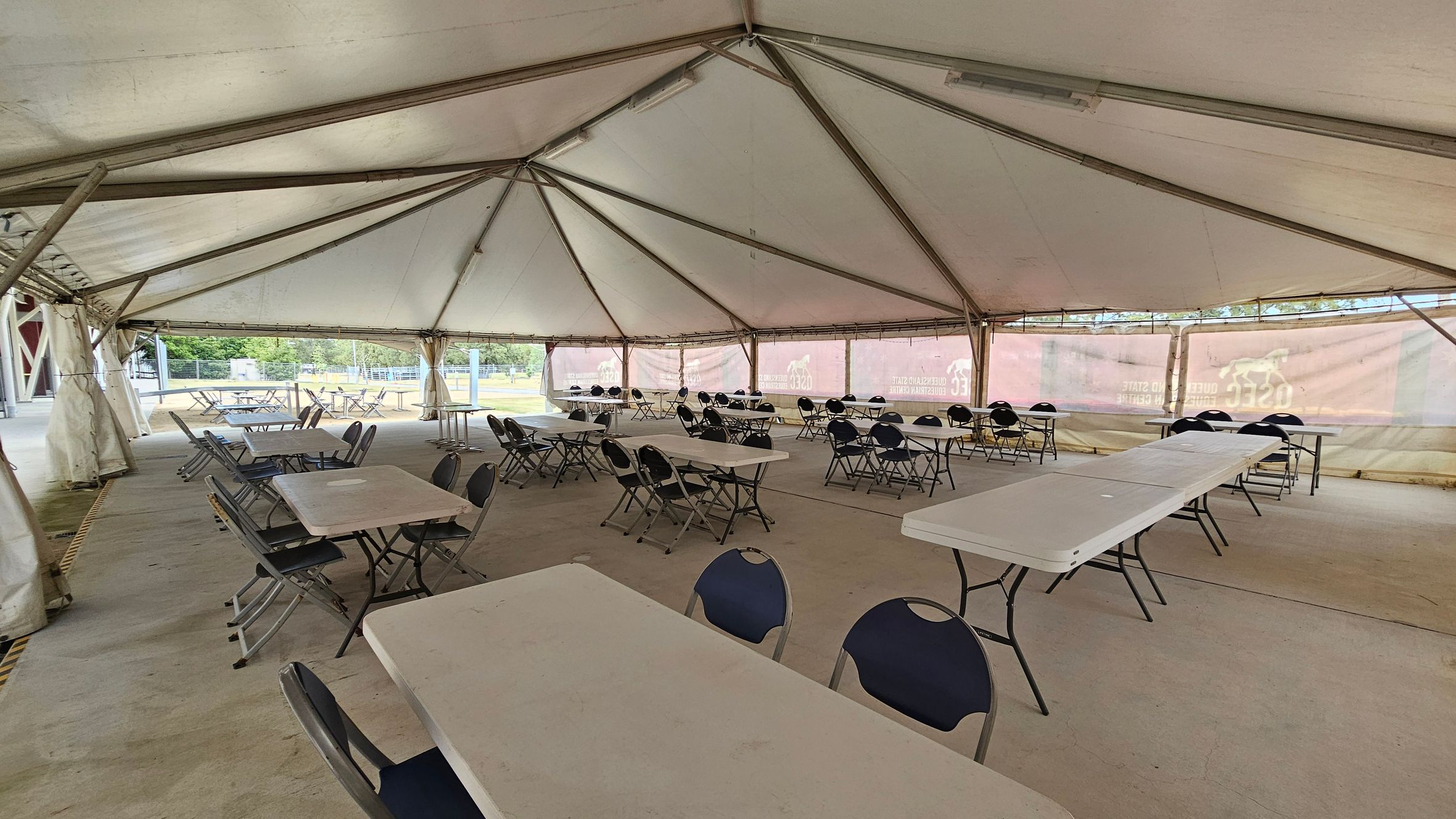 Inside a large tent with a high, triangular ceiling supported by metal beams. The floor is concrete. Several rectangular tables are arranged in rows, each surrounded by folding chairs. The tent walls are partially transparent, allowing a view of the outdoor area with greenery and structures in the background. The space appears set up for an event or gathering, with ample seating available.