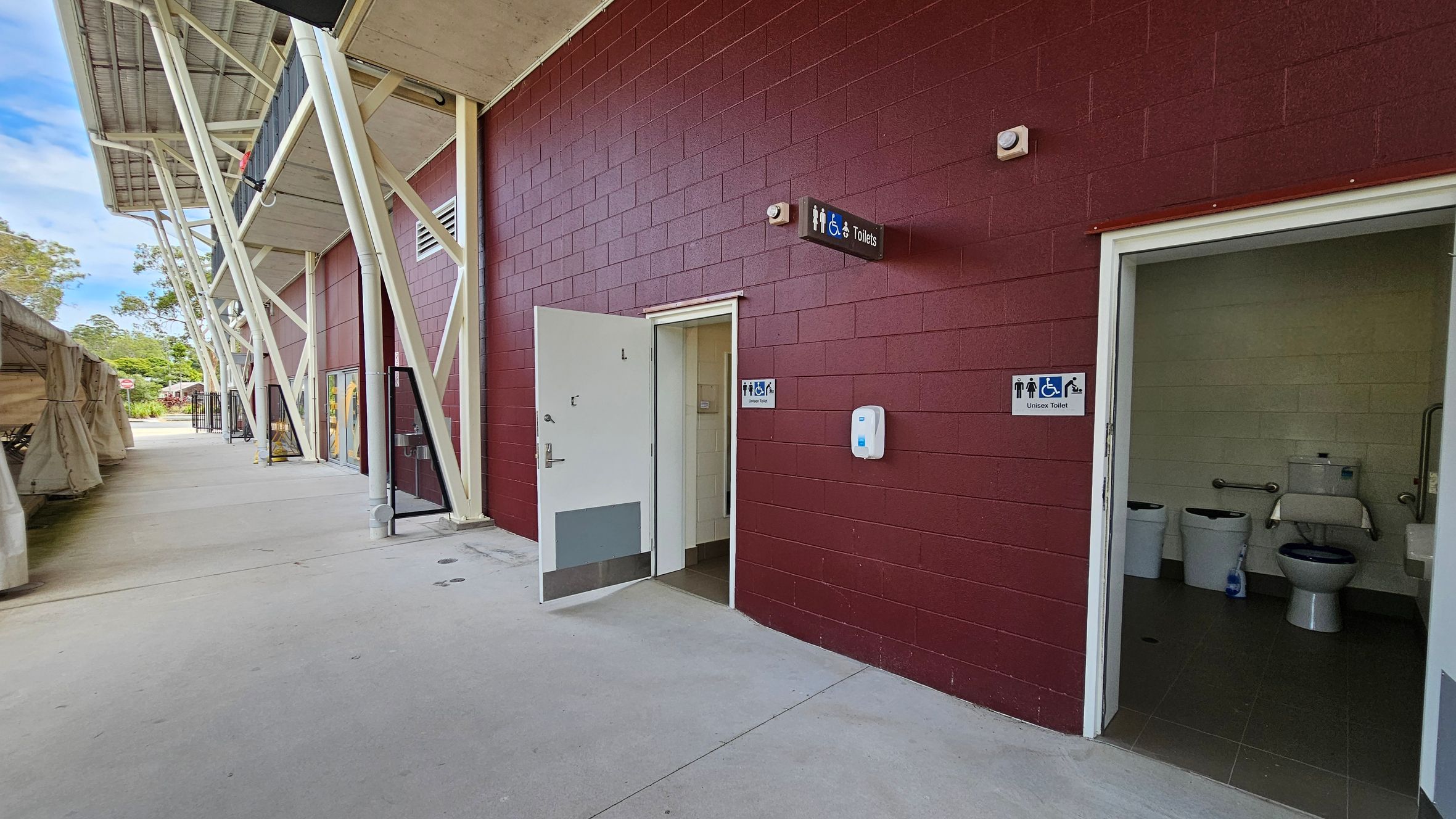 A red brick building with a unisex toilet entrance on the right. The door is open, revealing a toilet with grab bars and a sink. A sign above the door indicates accessibility features. On the left, a covered walkway with white structural beams and a concrete floor extends into the distance. A hand sanitiser dispenser is mounted on the wall near the entrance. Trees and a tent-like structure are visible in the background.