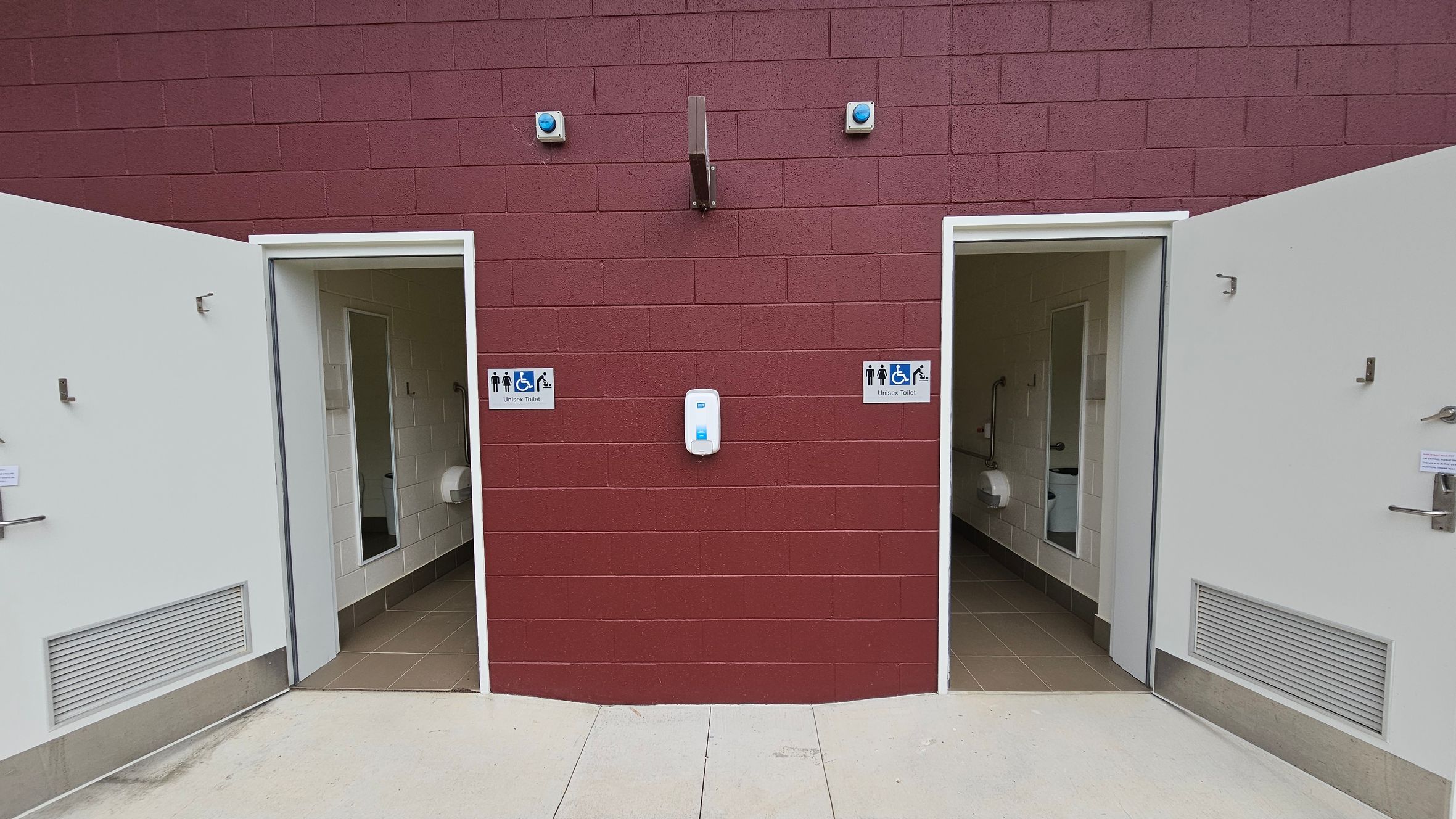 Two open doors on a maroon brick wall lead to unisex toilets, each marked with a sign showing accessibility symbols. Between the doors, a white dispenser is mounted on the wall. The interior of the toilets reveals white tiled walls and beige floors, with visible toilet paper dispensers. The floor outside is light grey concrete. Two small blue lights are above the doors, and a metal fixture is centred above the dispenser.