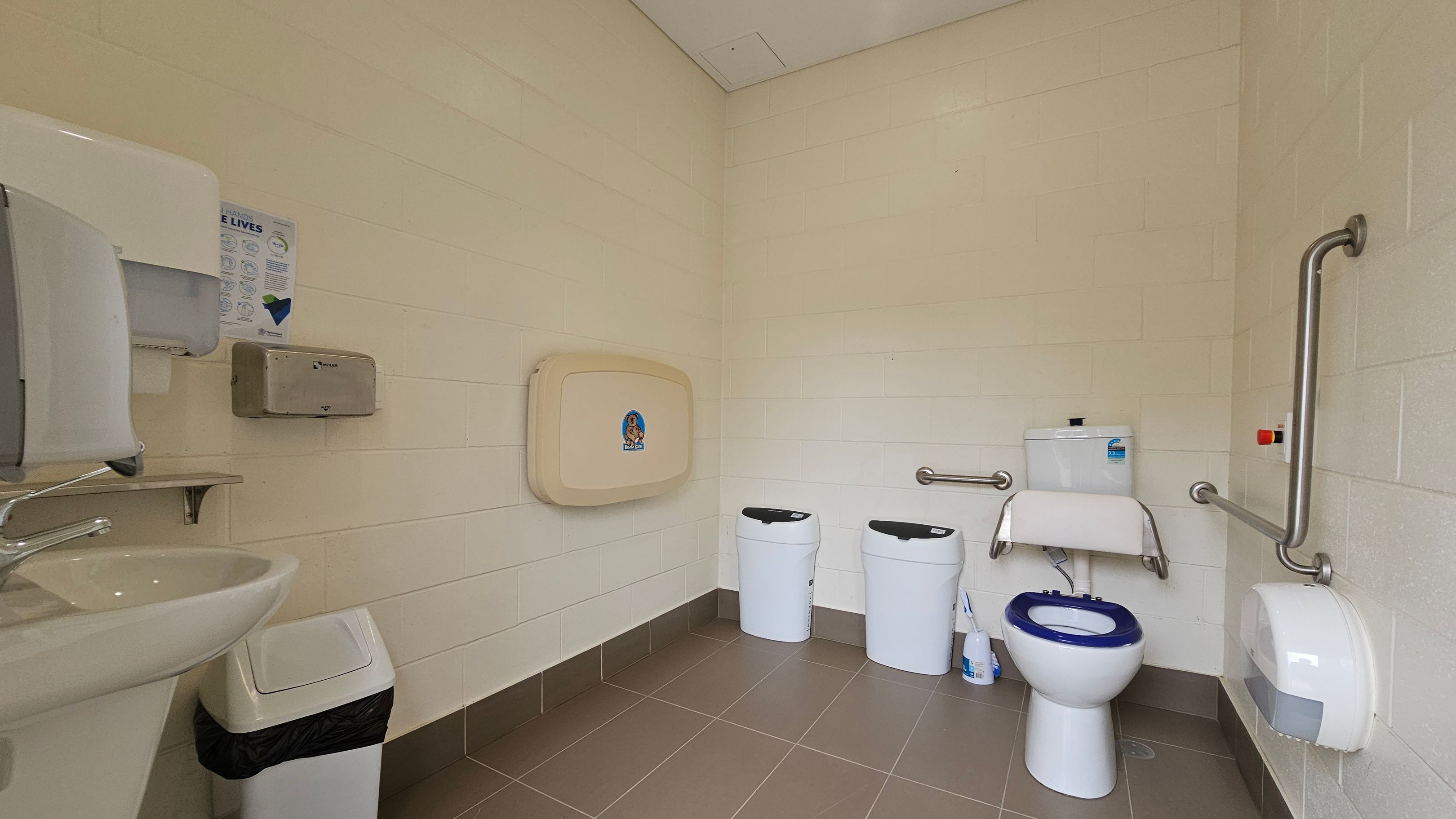 A public restroom with a beige tiled floor and cream walls. On the left, a sink with a soap dispenser and a paper towel holder above a small bin. In the centre, a baby changing station is mounted on the wall. On the right, a toilet with a blue seat, flanked by two white bins and a toilet paper dispenser. Stainless steel grab bars are installed around the toilet area. The ceiling is plain and light-coloured.