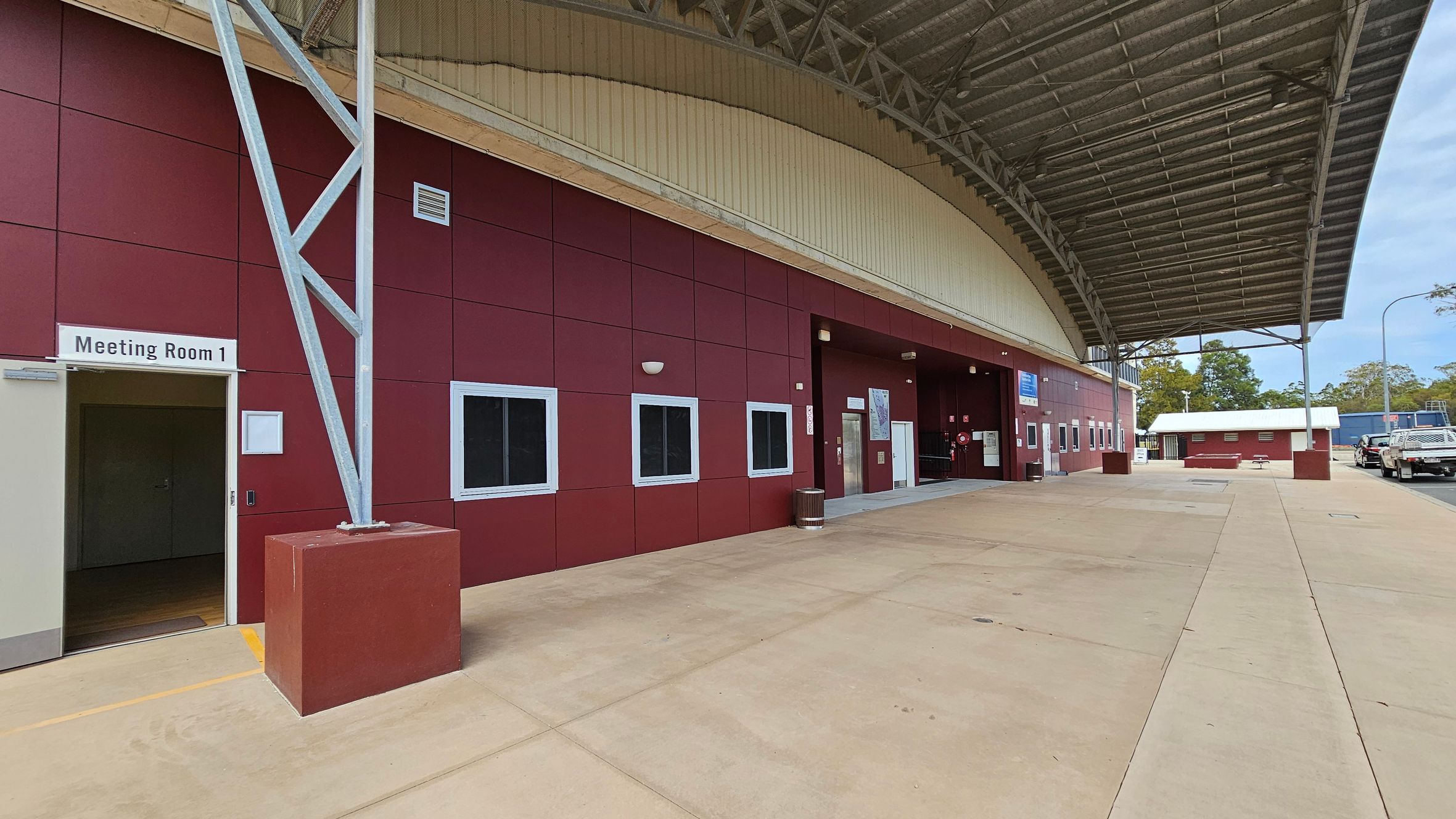 A large covered area with a red building on the left featuring several windows and a sign for "Meeting Room 1" above an open door. The structure has a metal support beam and a high, curved roof. The ground is paved with concrete. In the background, there are trees and a few parked vehicles on the right. A smaller red building is visible further back. The ceiling is made of metal with visible beams.