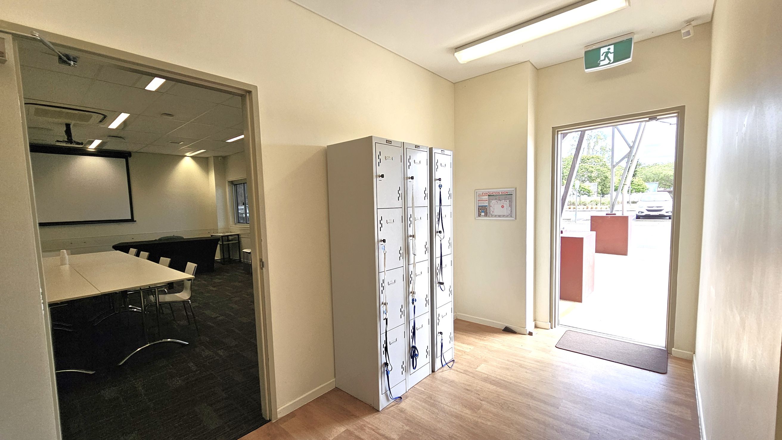 A hallway with light wooden flooring and cream walls. On the left, an open door leads to a room with a large screen, tables, and chairs. In the centre, a set of white lockers with lanyards hanging from them. On the right, a glass door opens to the outside, with a visible car and trees. Above the door, an exit sign is displayed. A small evacuation zone map is mounted on the wall near the lockers. The ceiling has fluorescent lights.