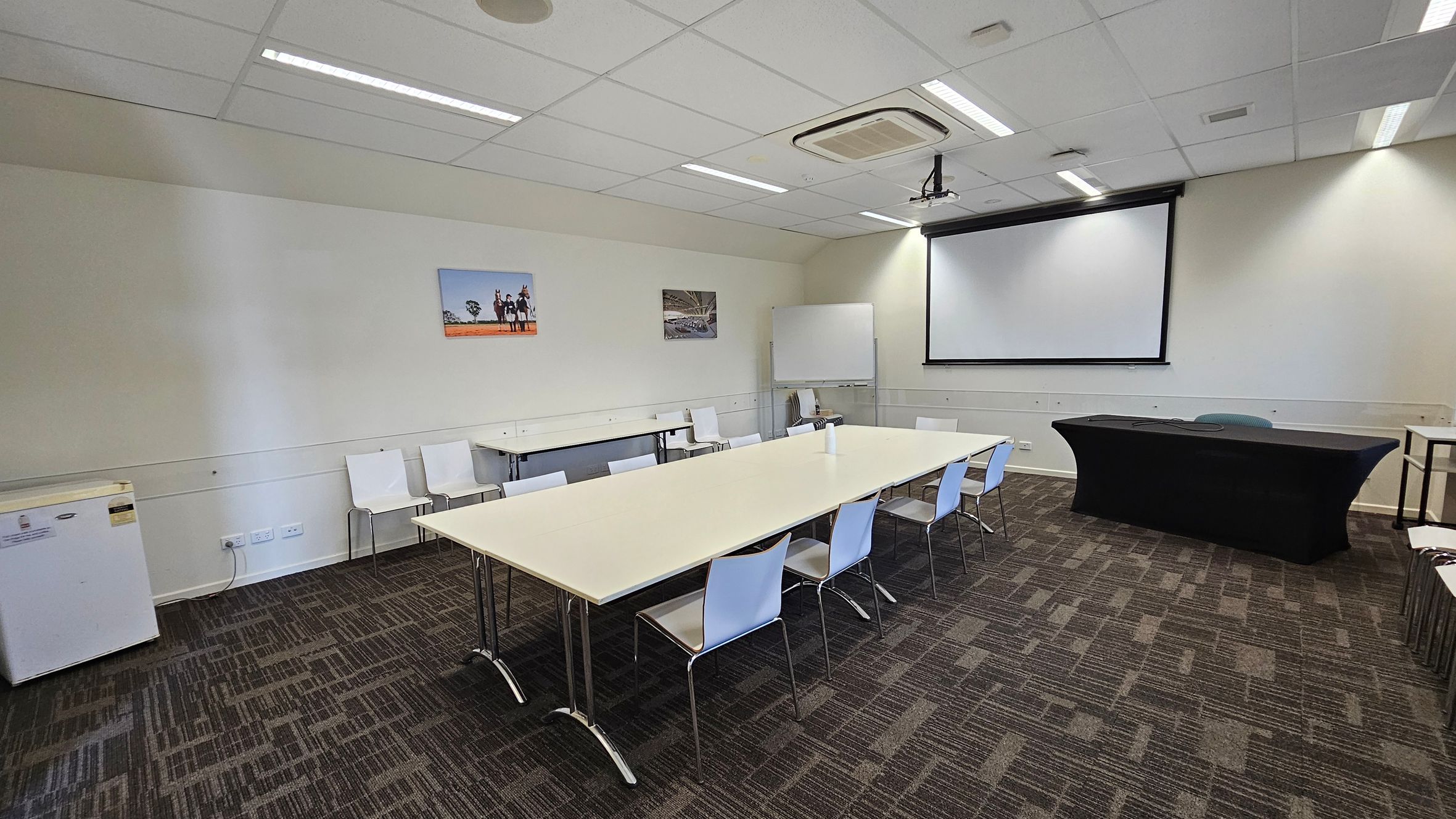 A conference room with a rectangular table in the centre surrounded by white chairs. On the right, there's a black table covered with a cloth and a large projection screen above it. A whiteboard is positioned against the back wall. The walls are light-coloured with two framed pictures. The floor is carpeted with a geometric pattern. The ceiling has recessed lighting and an air conditioning unit. A small fridge is on the left side.