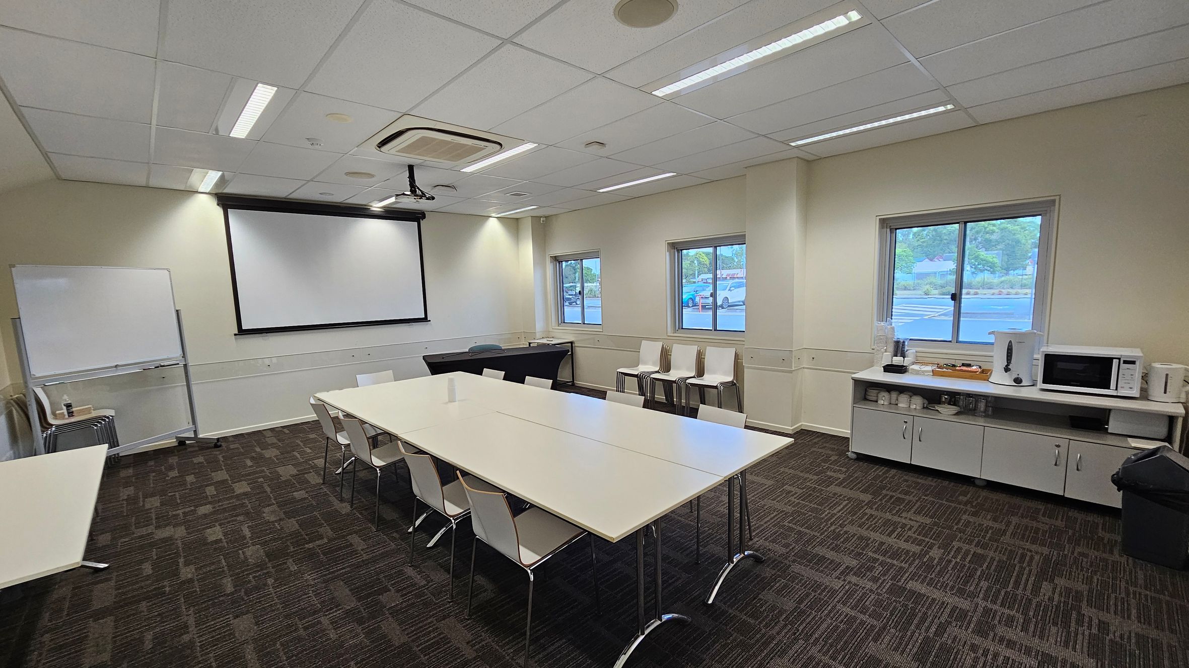 A conference room with a whiteboard and a large projection screen on the left wall. In the centre, there are several white tables arranged in a U-shape with white chairs around them. On the right, a counter with a microwave, cups, and a water dispenser is beneath two windows. The floor is carpeted with a dark, patterned design, and the ceiling has recessed lighting and an air conditioning unit.