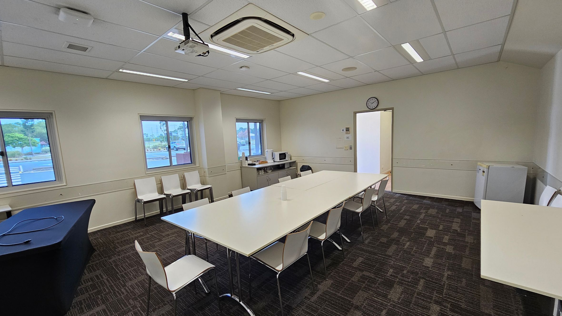 A meeting room with a large rectangular table in the centre surrounded by white chairs. On the left, there are two windows with a view outside and a table covered with a black cloth. The right side has a small fridge and a door leading to another room. The back wall features a clock and a counter with a microwave and other items. The ceiling has a projector and fluorescent lights, while the floor is carpeted with a dark pattern.