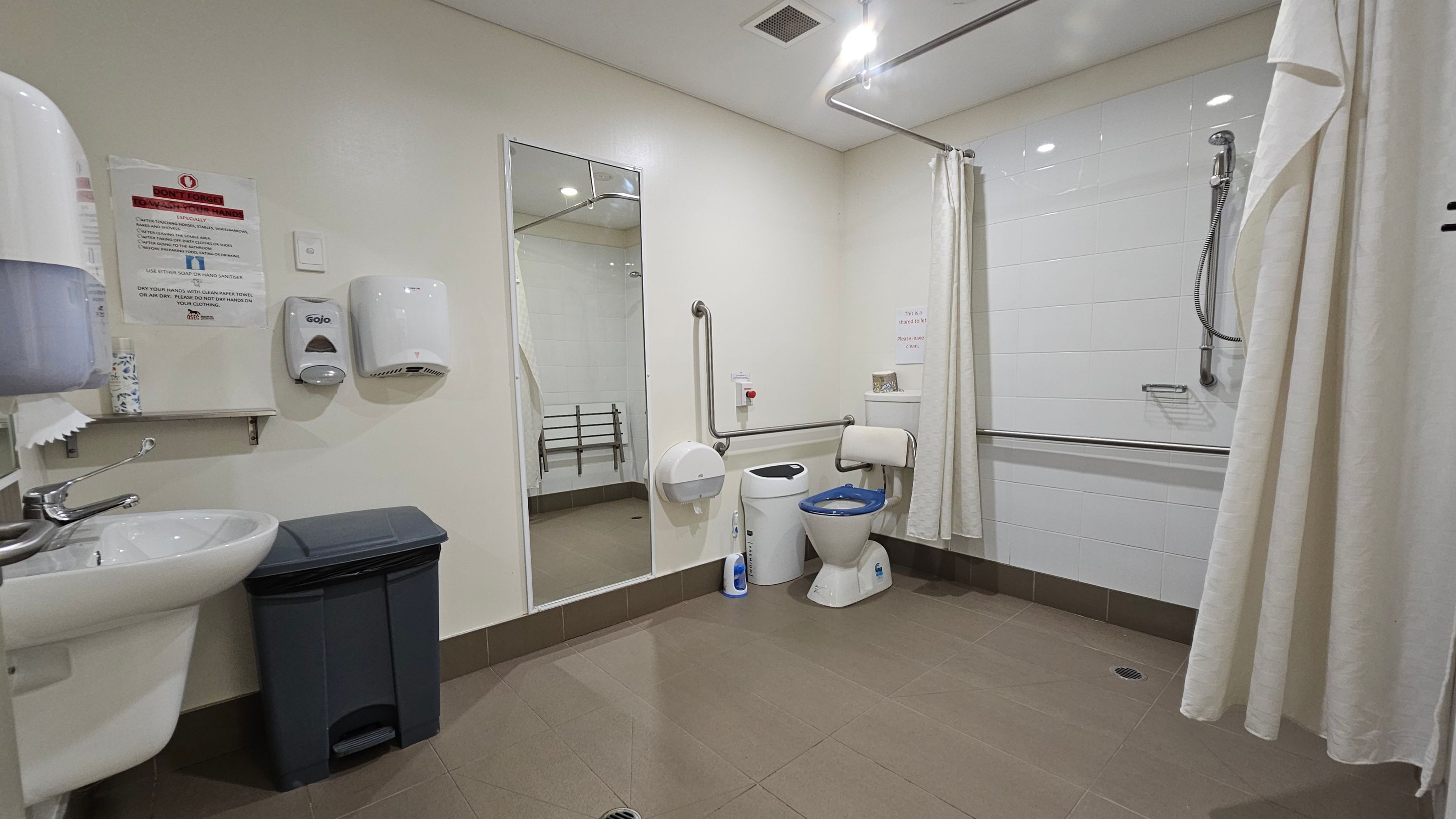 A spacious, accessible bathroom with a light-coloured floor and ceiling. On the left, a sink with a soap dispenser and a paper towel holder is mounted on the wall above a bin. A large mirror is centrally positioned, reflecting the room. On the right, a toilet with a blue seat is next to a wall-mounted grab bar. A shower area with a handheld showerhead and a curtain is visible, with additional grab bars for support. The walls are tiled in white.