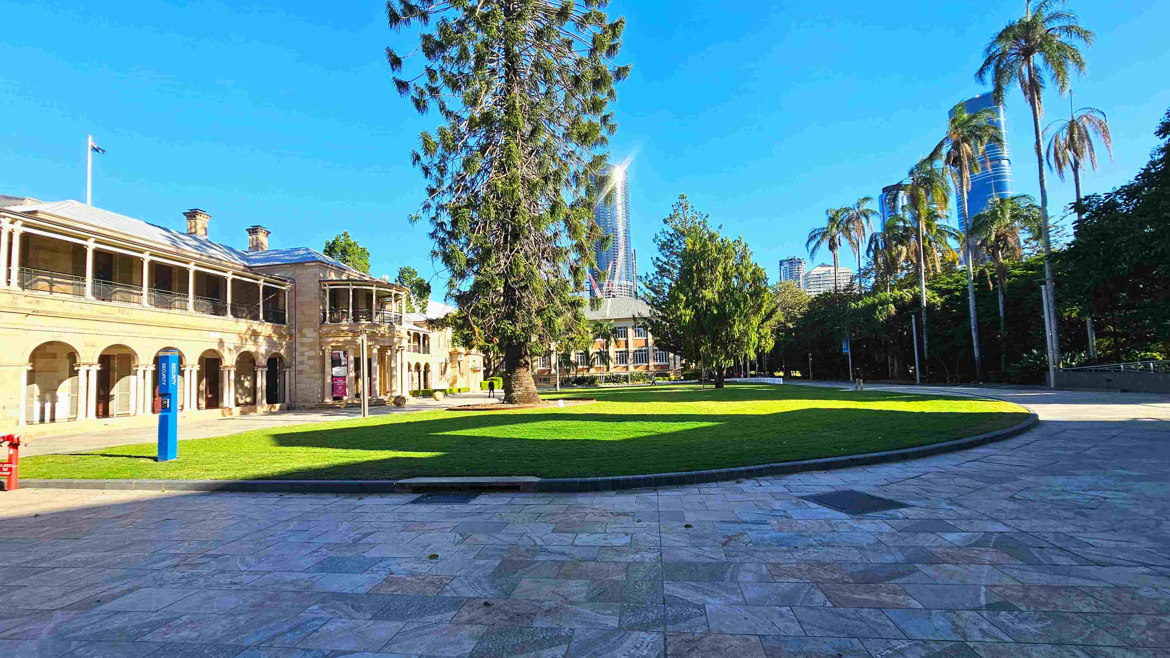 A spacious outdoor area with a large, well-maintained lawn bordered by a stone pathway. On the left, a historic building with arched windows and a flagpole stands prominently. Tall palm trees line the right side, and modern skyscrapers are visible in the background. A blue information sign is near the building, and the area feels open with a mix of natural and architectural elements. A spacious outdoor area with a large, well-maintained lawn bordered by a stone pathway. On the left, a historic building with arched windows and a flagpole stands prominently. Tall palm trees line the right side, and modern skyscrapers are visible in the background. A blue information sign is near the building, and the area feels open with a mix of natural and architectural elements.