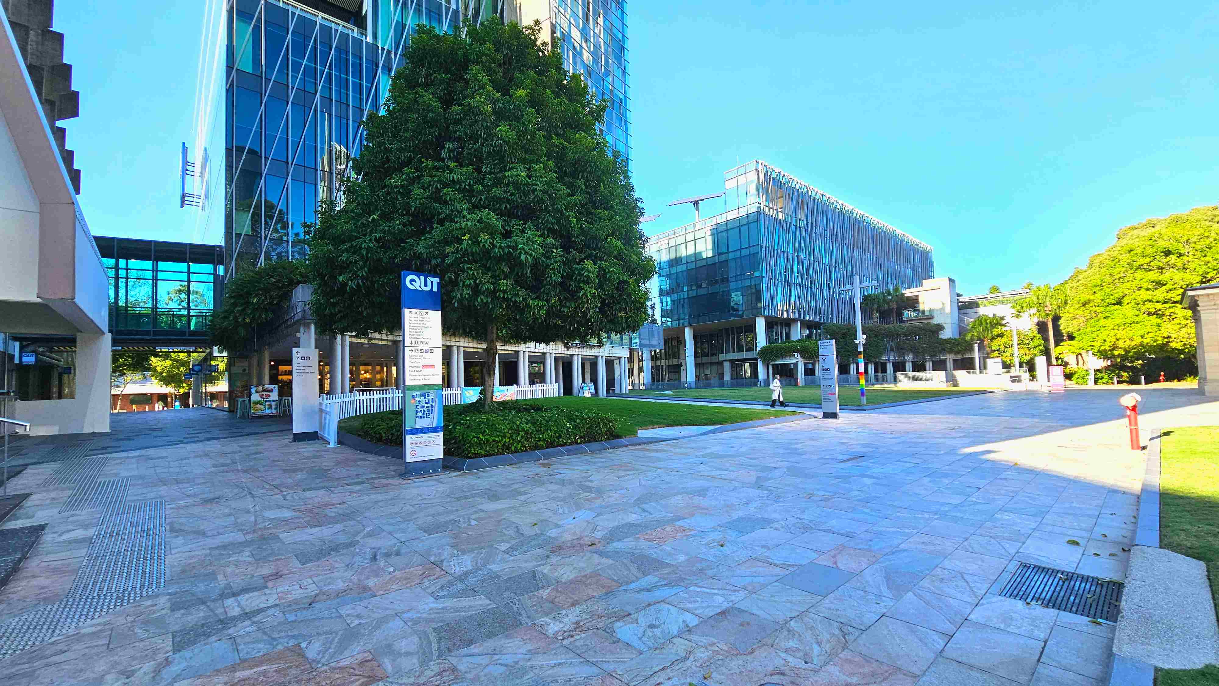 A modern urban plaza with a wide stone-tiled surface, bordered by a large glass building with a crisscross pattern and a smaller building with a similar design. A prominent sign with 'QUT' stands near a lush green tree, surrounded by low hedges. A pedestrian walkway with tactile paving runs alongside. The area is open and spacious, with a few people walking and a red fire hydrant visible on the right. A modern urban plaza with a wide stone-tiled surface, bordered by a large glass building with a crisscross pattern and a smaller building with a similar design. A prominent sign with 'QUT' stands near a lush green tree, surrounded by low hedges. A pedestrian walkway with tactile paving runs alongside. The area is open and spacious, with a few people walking and a red fire hydrant visible on the right.