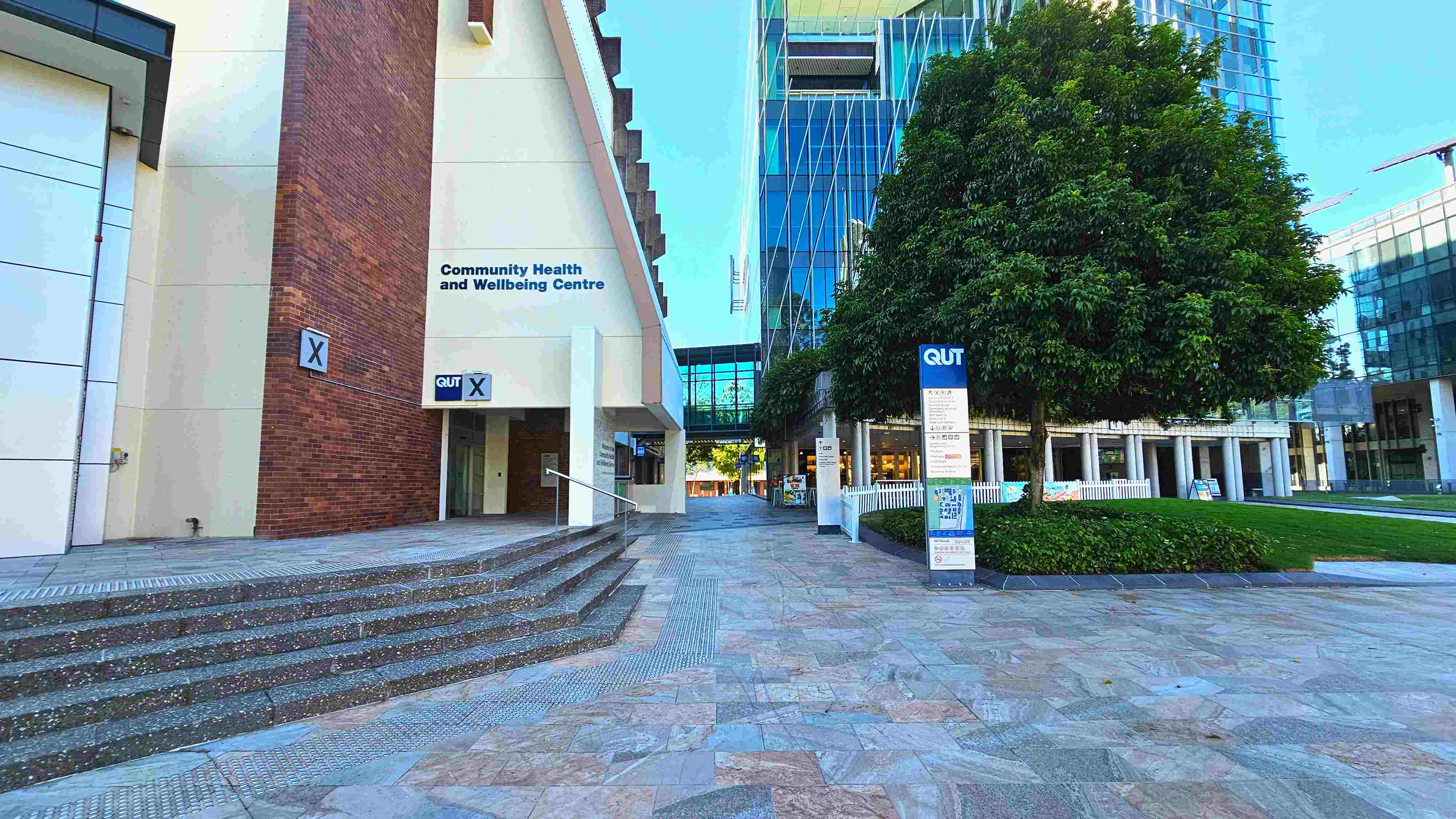 A modern urban space featuring a Community Health and Wellbeing Centre with a brick and cream facade on the left. A set of wide, shallow steps leads up to the entrance. To the right, a large tree stands beside a signpost with 'QUT' branding. The ground is paved with patterned stone tiles. In the background, a glass-fronted building with a covered walkway connects the structures, creating a semi-enclosed area. A modern urban space featuring a Community Health and Wellbeing Centre with a brick and cream facade on the left. A set of wide, shallow steps leads up to the entrance. To the right, a large tree stands beside a signpost with 'QUT' branding. The ground is paved with patterned stone tiles. In the background, a glass-fronted building with a covered walkway connects the structures, creating a semi-enclosed area.