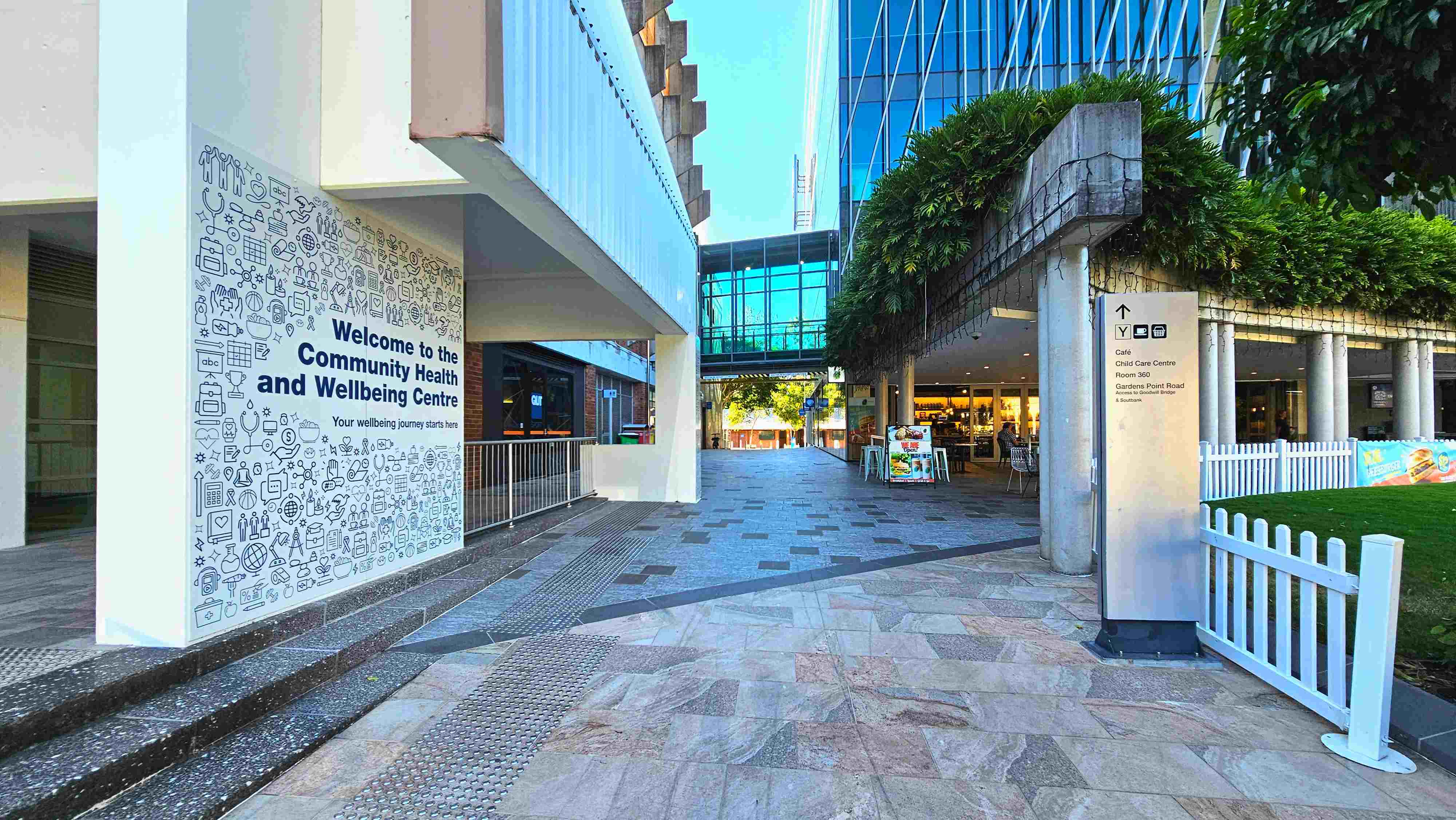 A modern outdoor walkway with a large sign reading 'Welcome to the Community Health and Wellbeing Centre' on a wall to the left. The path is paved with a mix of grey and pink tiles, leading towards a glass-walled building in the background. A white fence borders a grassy area on the right, and a tall signpost provides directions to various facilities. Greenery hangs from a concrete overhang, adding a touch of nature to the urban setting. A modern outdoor walkway with a large sign reading 'Welcome to the Community Health and Wellbeing Centre' on a wall to the left. The path is paved with a mix of grey and pink tiles, leading towards a glass-walled building in the background. A white fence borders a grassy area on the right, and a tall signpost provides directions to various facilities. Greenery hangs from a concrete overhang, adding a touch of nature to the urban setting.