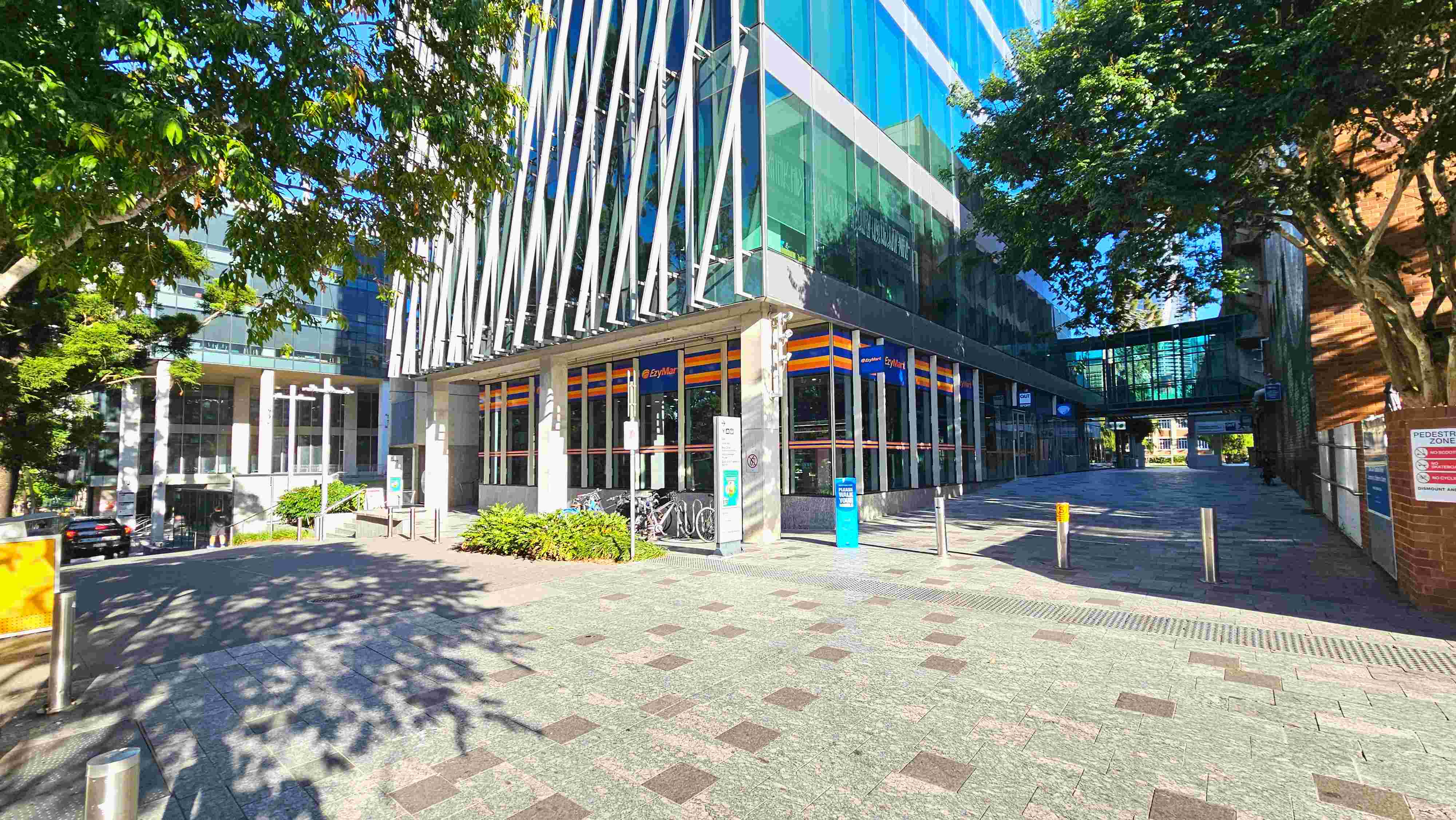 A modern urban scene featuring a glass-fronted building with diagonal white beams and a convenience store on the ground floor. The pavement is a mix of grey and brown tiles, leading to a pedestrian zone marked by bollards. A tree provides shade on the left, and a glass skybridge connects buildings in the background. Bicycles are parked near the entrance, and signage is visible on the walls. A modern urban scene featuring a glass-fronted building with diagonal white beams and a convenience store on the ground floor. The pavement is a mix of grey and brown tiles, leading to a pedestrian zone marked by bollards. A tree provides shade on the left, and a glass skybridge connects buildings in the background. Bicycles are parked near the entrance, and signage is visible on the walls.