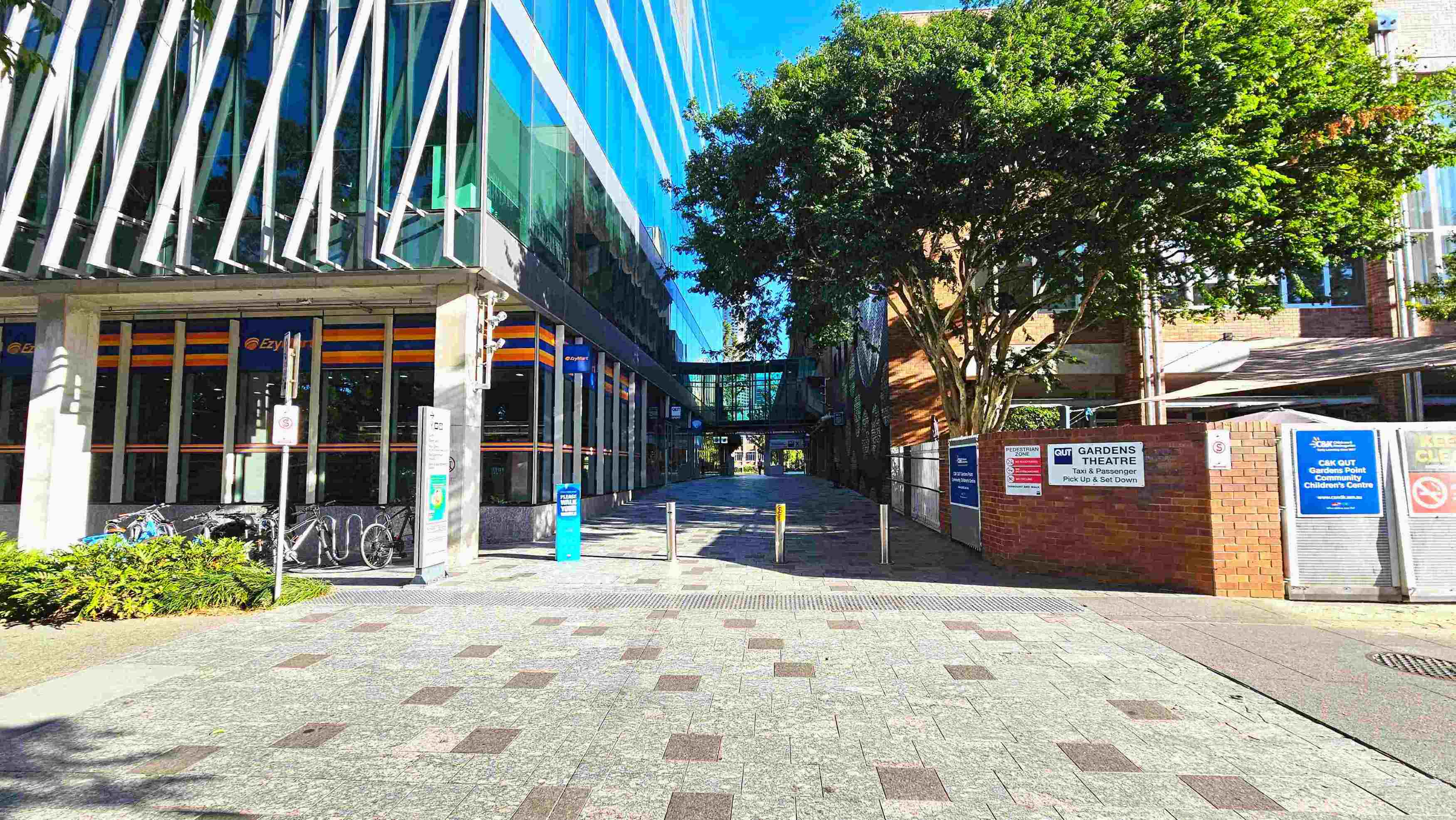 A modern urban walkway flanked by a glass-fronted building on the left and a brick building on the right. The glass building features diagonal white beams and a row of bicycles parked outside. A tree provides shade near the brick building, which has signage for Gardens Theatre. The path is paved with grey and brown tiles, leading to a glass bridge connecting the buildings. Bollards mark the entrance to the walkway. A modern urban walkway flanked by a glass-fronted building on the left and a brick building on the right. The glass building features diagonal white beams and a row of bicycles parked outside. A tree provides shade near the brick building, which has signage for Gardens Theatre. The path is paved with grey and brown tiles, leading to a glass bridge connecting the buildings. Bollards mark the entrance to the walkway.