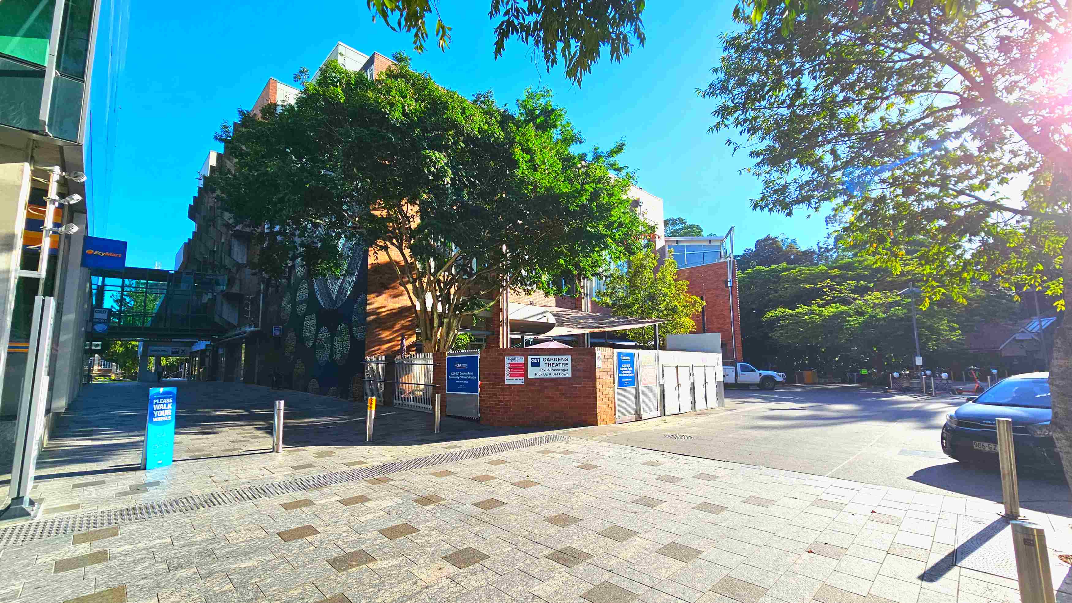 A paved area with a mix of light and dark grey tiles leads to a brick building partially obscured by lush green trees. Signs on the building indicate it is the QUT Gardens Theatre. A glass walkway connects to another structure on the left. Metal bollards line the path, and a parked car is visible on the right. The area feels open with trees providing shade and a clear view of the buildings. A paved area with a mix of light and dark grey tiles leads to a brick building partially obscured by lush green trees. Signs on the building indicate it is the QUT Gardens Theatre. A glass walkway connects to another structure on the left. Metal bollards line the path, and a parked car is visible on the right. The area feels open with trees providing shade and a clear view of the buildings.