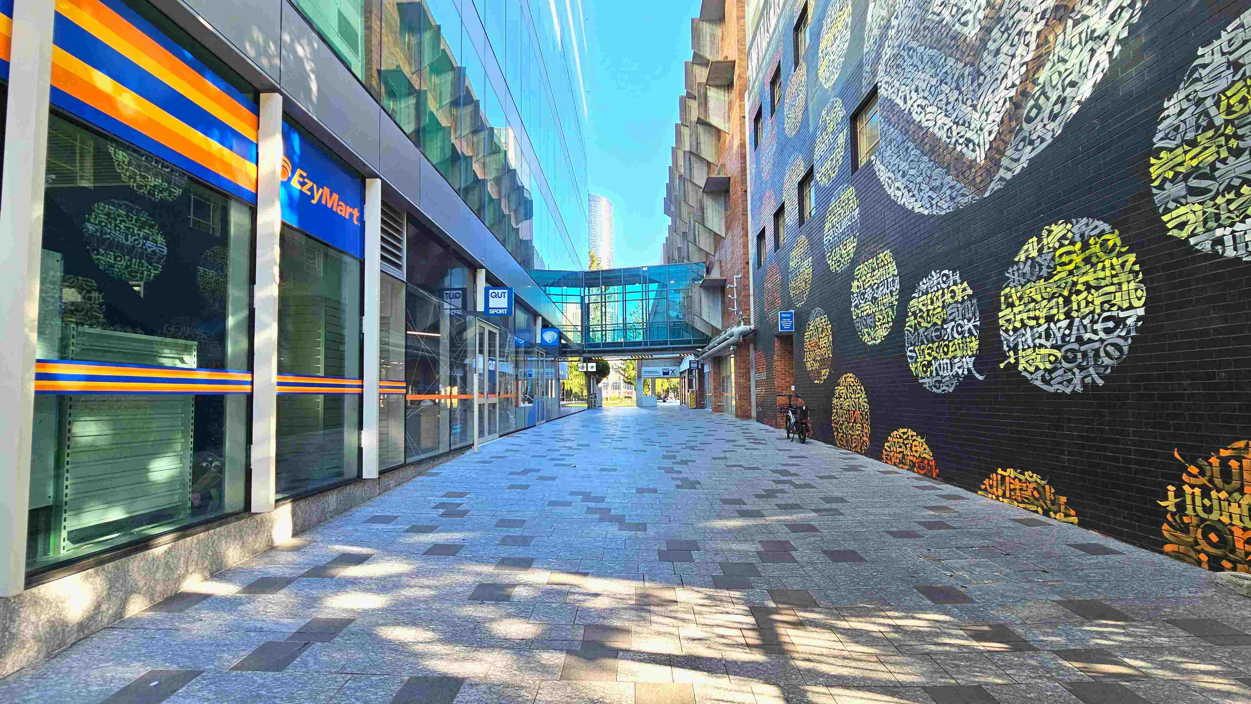 A pedestrian walkway flanked by a modern building with glass windows on the left and a brick wall adorned with colourful circular graffiti on the right. The path is paved with grey and brown tiles. A glass skybridge connects the buildings above. A bicycle is parked against the brick wall. Signs for 'EzyMart' and 'QUT Sport' are visible on the left. The area appears open and well-lit. A pedestrian walkway flanked by a modern building with glass windows on the left and a brick wall adorned with colourful circular graffiti on the right. The path is paved with grey and brown tiles. A glass skybridge connects the buildings above. A bicycle is parked against the brick wall. Signs for 'EzyMart' and 'QUT Sport' are visible on the left. The area appears open and well-lit.