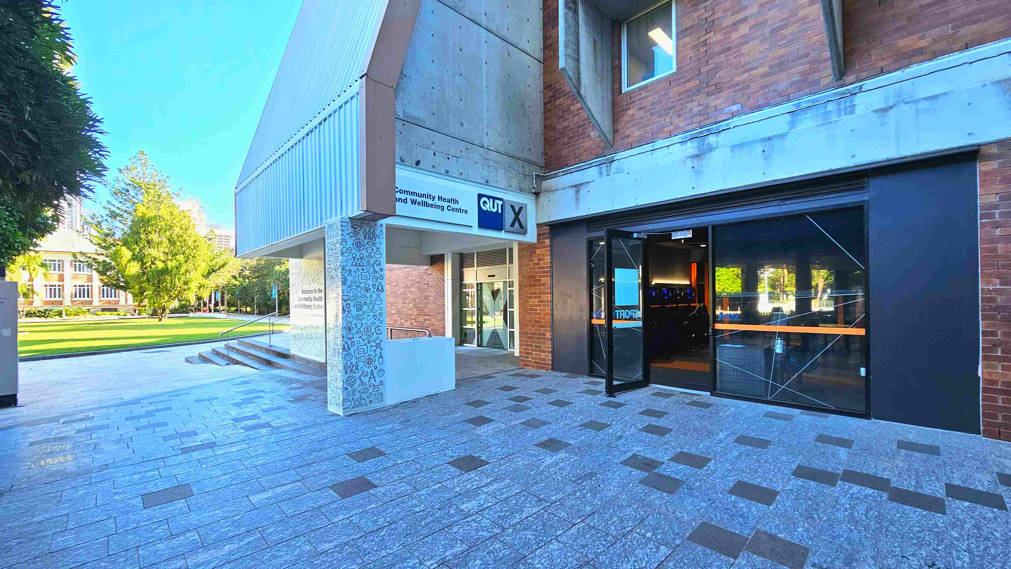 The image shows the entrance to a building with a sign reading 'Community Health and Wellbeing Centre' and 'QUT X'. The structure features a mix of brick and concrete with a modern design. A glass door is open, revealing a dimly lit interior. The ground is paved with grey tiles interspersed with darker squares. To the left, a grassy area with trees and a historic building is visible, suggesting an open campus environment. The image shows the entrance to a building with a sign reading 'Community Health and Wellbeing Centre' and 'QUT X'. The structure features a mix of brick and concrete with a modern design. A glass door is open, revealing a dimly lit interior. The ground is paved with grey tiles interspersed with darker squares. To the left, a grassy area with trees and a historic building is visible, suggesting an open campus environment.