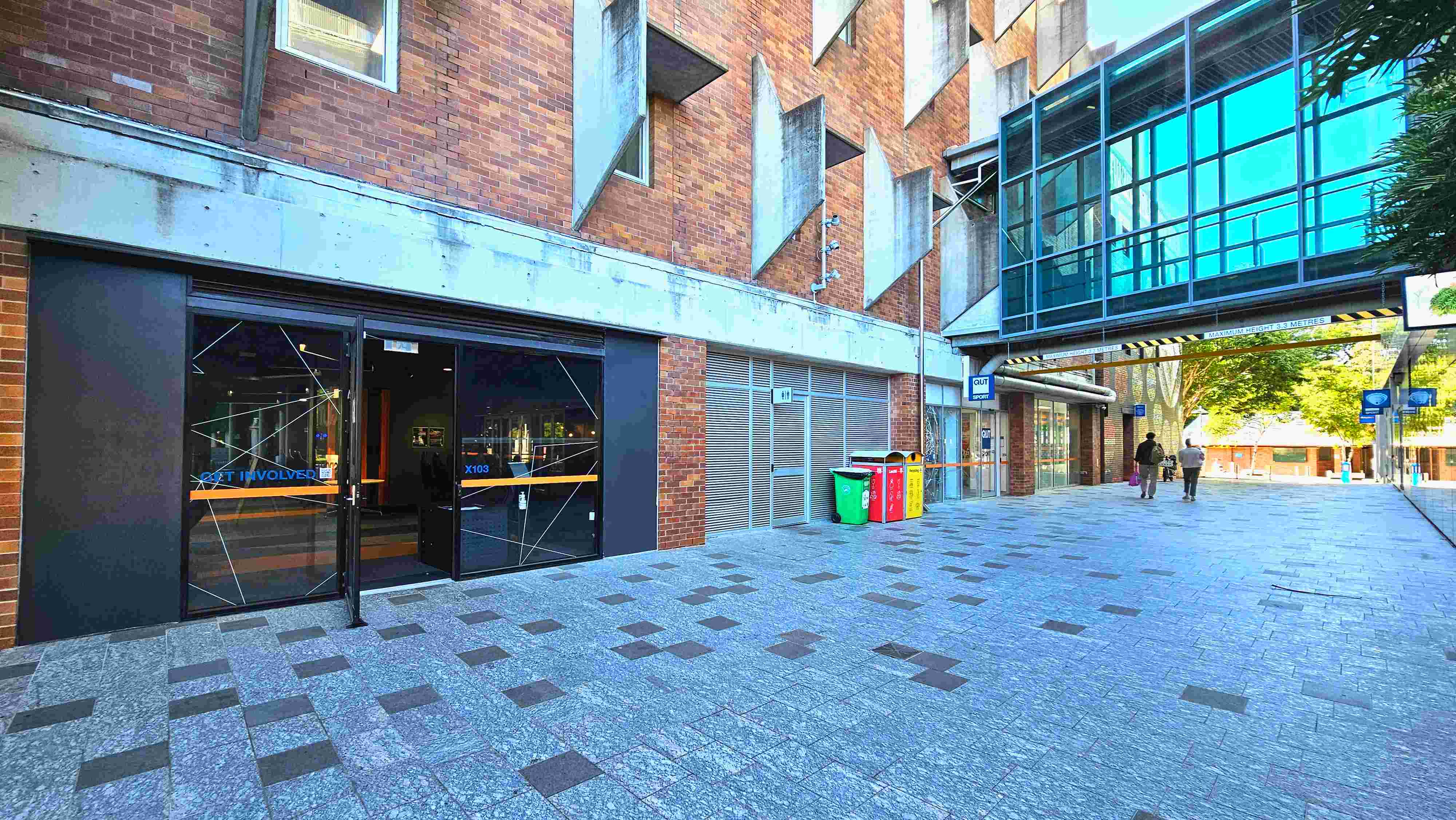 A modern urban walkway with a brick building on the left featuring geometric concrete protrusions. The building has a glass entrance with 'Get Involved' signage. Adjacent are colourful recycling bins. A glass skybridge connects to another structure, casting reflections on the ground. The walkway is paved with grey tiles interspersed with darker squares, leading to a tree-lined area in the distance. A modern urban walkway with a brick building on the left featuring geometric concrete protrusions. The building has a glass entrance with 'Get Involved' signage. Adjacent are colourful recycling bins. A glass skybridge connects to another structure, casting reflections on the ground. The walkway is paved with grey tiles interspersed with darker squares, leading to a tree-lined area in the distance.