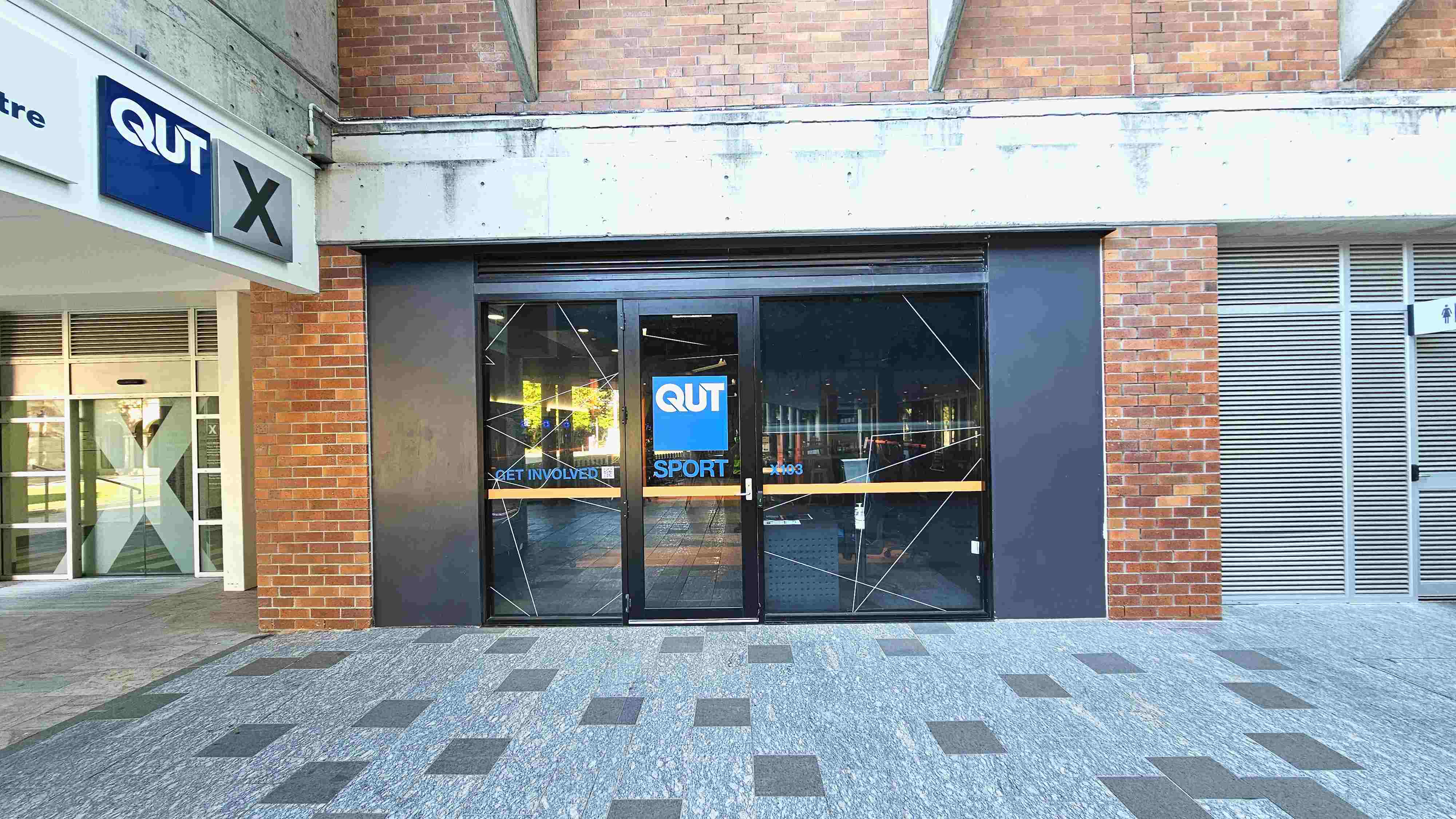 The image shows a brick building facade with a central glass entrance featuring the QUT Sport logo. The entrance has two large glass doors with geometric patterns and a blue QUT sign. To the left, a sign reads 'QUT X' above a glass door. The ground is paved with grey tiles in a checkered pattern. On the right, there is a louvred panel and a small sign indicating restrooms. The image shows a brick building facade with a central glass entrance featuring the QUT Sport logo. The entrance has two large glass doors with geometric patterns and a blue QUT sign. To the left, a sign reads 'QUT X' above a glass door. The ground is paved with grey tiles in a checkered pattern. On the right, there is a louvred panel and a small sign indicating restrooms.