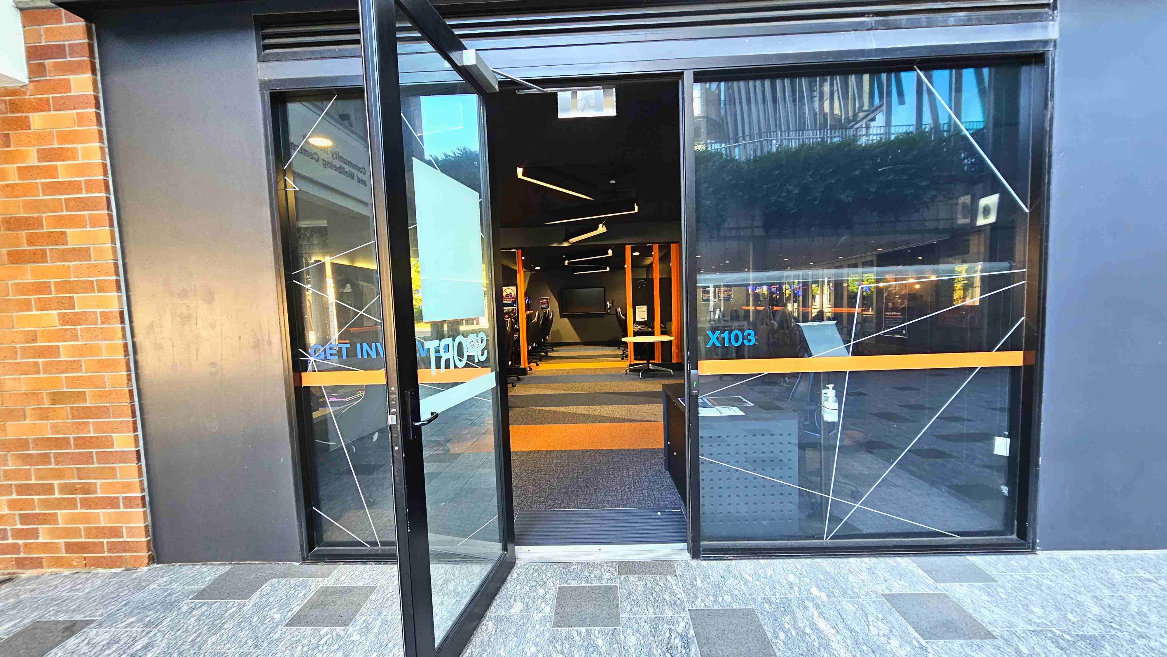 A modern entrance with large glass doors, one of which is open, revealing an interior with a patterned carpet and sleek black and orange furnishings. The exterior features a brick wall on the left and a dark grey panel on the right. The floor outside is tiled with grey and black squares. Inside, linear ceiling lights and a row of chairs are visible. A hand sanitiser dispenser is placed near the entrance. A modern entrance with large glass doors, one of which is open, revealing an interior with a patterned carpet and sleek black and orange furnishings. The exterior features a brick wall on the left and a dark grey panel on the right. The floor outside is tiled with grey and black squares. Inside, linear ceiling lights and a row of chairs are visible. A hand sanitiser dispenser is placed near the entrance.