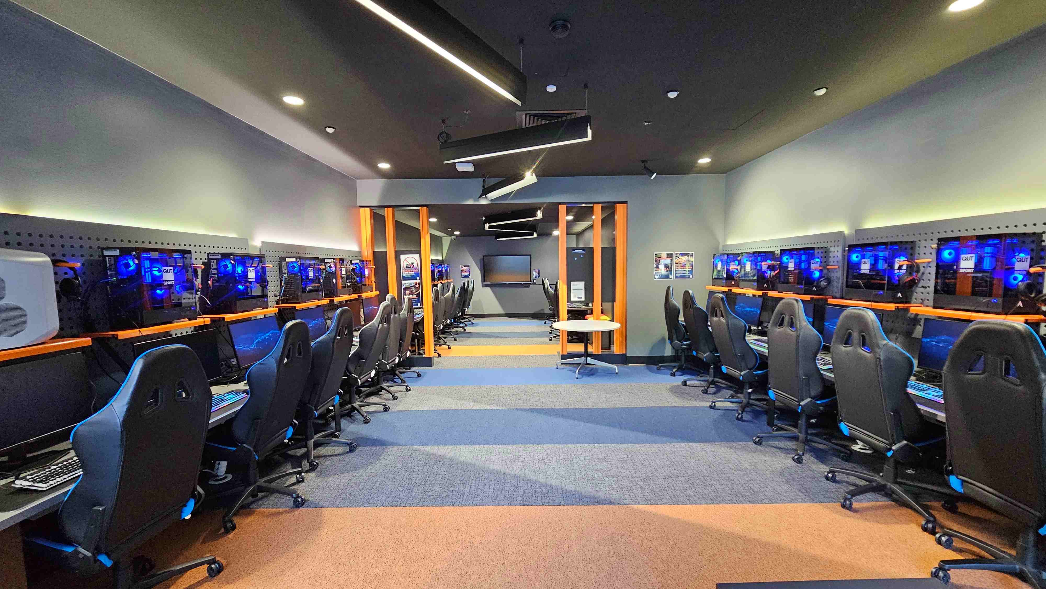 A modern gaming room with two rows of high-backed gaming chairs facing computer stations along both walls. Each station features a monitor and a glowing blue-lit computer tower mounted on a perforated panel. The room has a striped carpet in shades of blue and orange, and overhead, angular black light fixtures hang from the ceiling. At the far end, a large screen is mounted on the wall, flanked by orange vertical beams. A modern gaming room with two rows of high-backed gaming chairs facing computer stations along both walls. Each station features a monitor and a glowing blue-lit computer tower mounted on a perforated panel. The room has a striped carpet in shades of blue and orange, and overhead, angular black light fixtures hang from the ceiling. At the far end, a large screen is mounted on the wall, flanked by orange vertical beams.