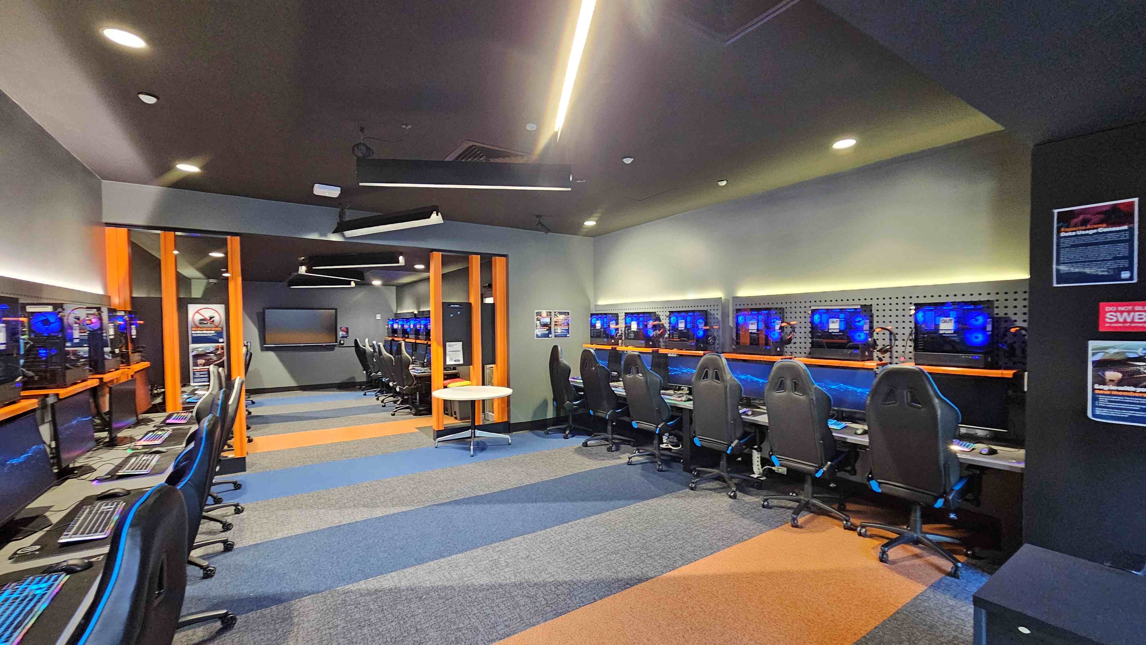 A modern gaming room with rows of high-backed gaming chairs facing computer stations. Each station has a monitor with blue LED lighting. The room features a mix of grey, blue, and orange carpet tiles, creating a vibrant pattern. Orange vertical beams divide the space, and posters are visible on the walls. The ceiling has recessed lighting and angular fixtures, adding a futuristic touch. A modern gaming room with rows of high-backed gaming chairs facing computer stations. Each station has a monitor with blue LED lighting. The room features a mix of grey, blue, and orange carpet tiles, creating a vibrant pattern. Orange vertical beams divide the space, and posters are visible on the walls. The ceiling has recessed lighting and angular fixtures, adding a futuristic touch.