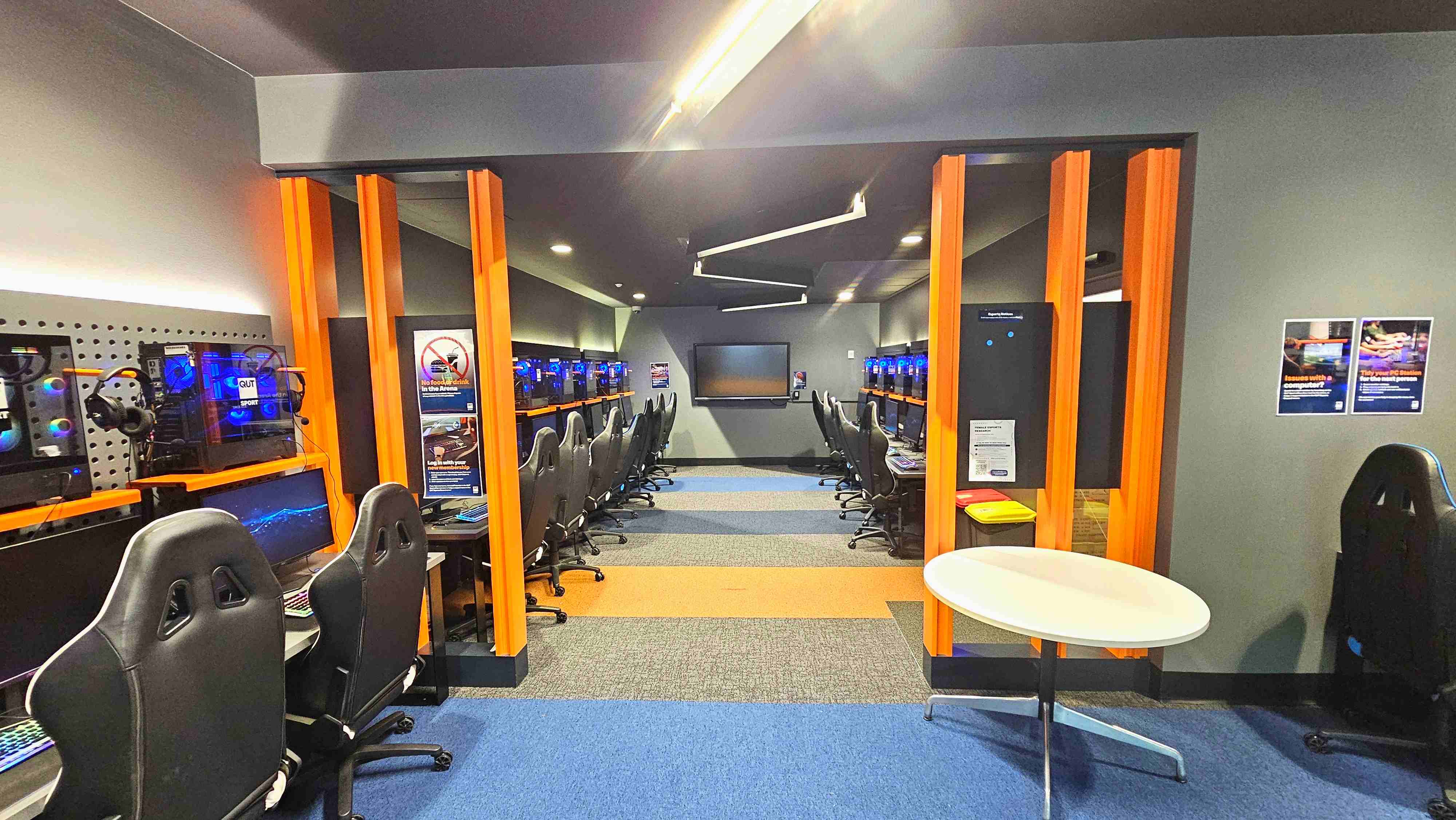 A modern gaming room with rows of high-backed black chairs facing computer stations on both sides. Each station has a monitor, keyboard, and headset, with blue-lit computer towers visible. The room is divided by orange vertical beams and features a grey wall with a large screen at the far end. The floor is carpeted in blue, orange, and grey sections. Posters are on the walls, and a small round table is in the foreground. A modern gaming room with rows of high-backed black chairs facing computer stations on both sides. Each station has a monitor, keyboard, and headset, with blue-lit computer towers visible. The room is divided by orange vertical beams and features a grey wall with a large screen at the far end. The floor is carpeted in blue, orange, and grey sections. Posters are on the walls, and a small round table is in the foreground.