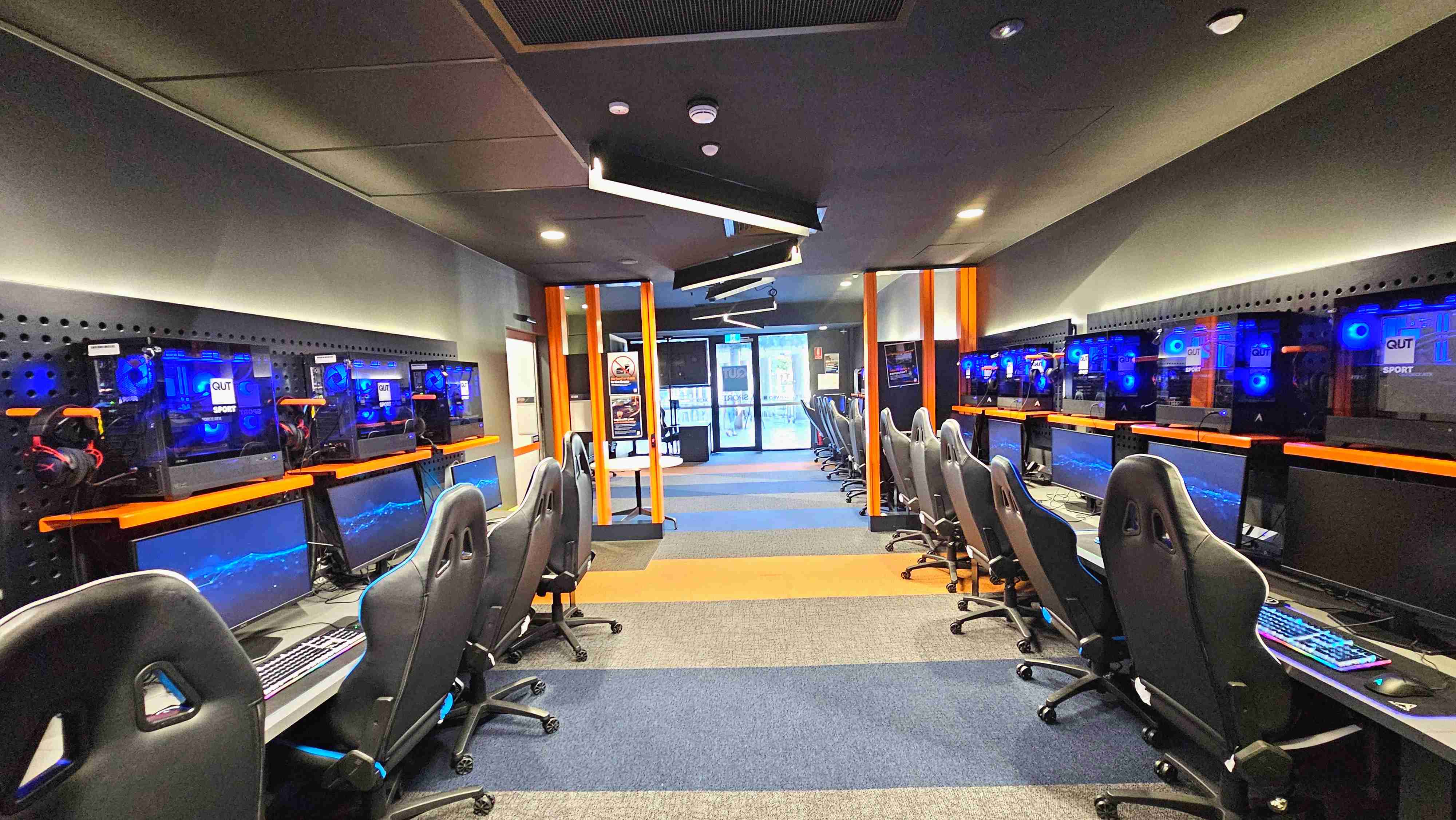 A modern gaming room with two rows of high-backed gaming chairs facing computer stations. Each station has a monitor, keyboard, and a glowing blue-lit CPU. The room features a dark ceiling with angular light fixtures and orange vertical beams. The floor is carpeted with alternating blue and orange stripes. At the far end, glass doors lead to another area, flanked by posters and signage. A modern gaming room with two rows of high-backed gaming chairs facing computer stations. Each station has a monitor, keyboard, and a glowing blue-lit CPU. The room features a dark ceiling with angular light fixtures and orange vertical beams. The floor is carpeted with alternating blue and orange stripes. At the far end, glass doors lead to another area, flanked by posters and signage.
