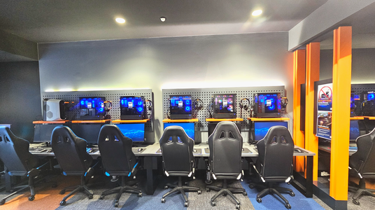 A row of five gaming stations is set against a wall with pegboard panels. Each station features a desktop computer with illuminated components, a monitor, keyboard, and headphones. Black gaming chairs are positioned in front of each setup. The floor is carpeted with a mix of grey and blue tiles. Bright orange vertical beams frame the right side, where a sign prohibits food and drinks. Ceiling lights provide illumination. A row of five gaming stations is set against a wall with pegboard panels. Each station features a desktop computer with illuminated components, a monitor, keyboard, and headphones. Black gaming chairs are positioned in front of each setup. The floor is carpeted with a mix of grey and blue tiles. Bright orange vertical beams frame the right side, where a sign prohibits food and drinks. Ceiling lights provide illumination.
