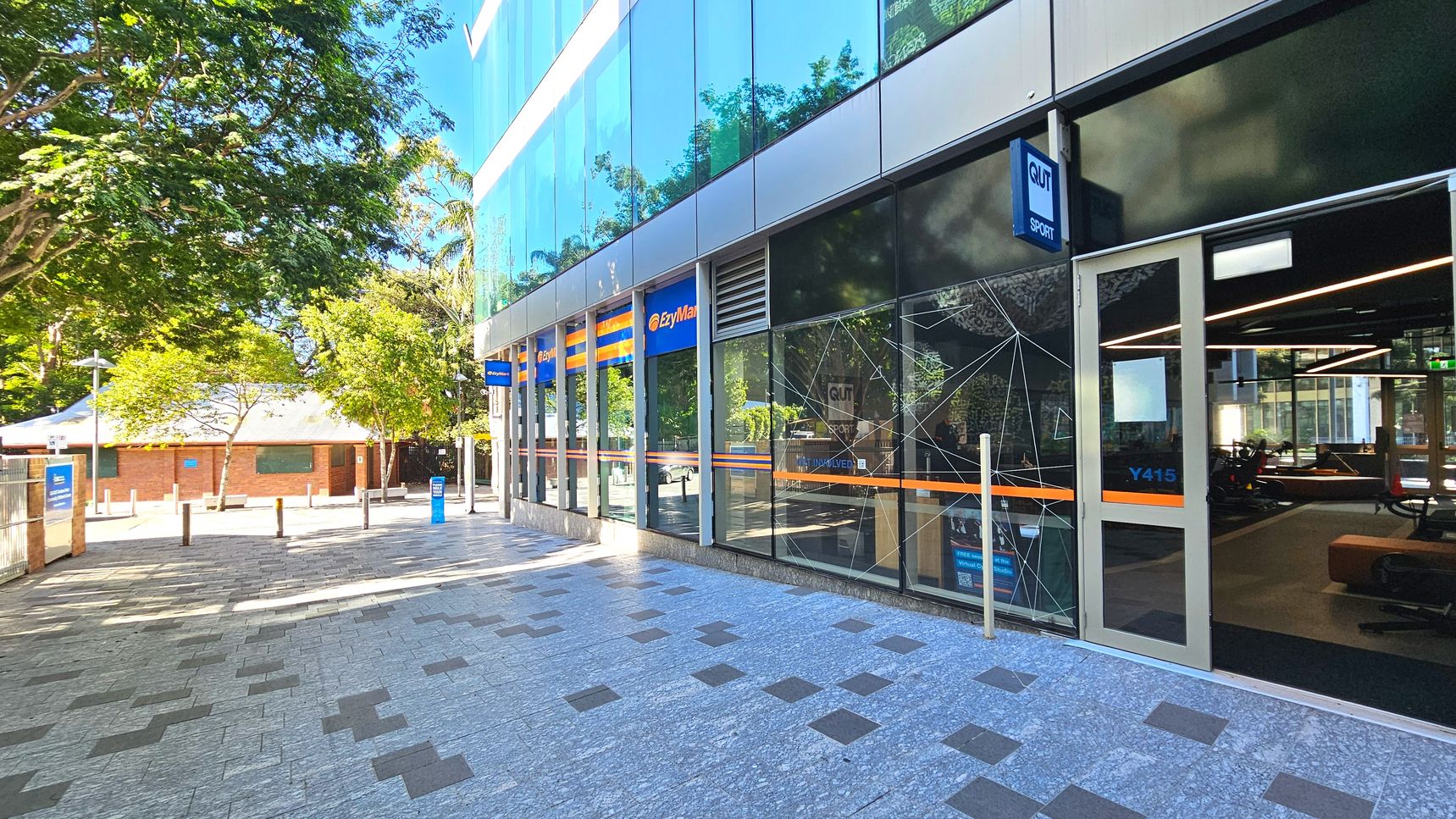 A modern building with large glass windows and a sign reading 'QUT Sport' above a door marked 'Y415'. The building's facade features blue and orange signage for 'EzyMart'. The pavement is a mix of grey and dark tiles, leading to a tree-lined area with bollards. A brick building with a white roof is visible in the background. Inside, gym equipment is visible through the glass. A modern building with large glass windows and a sign reading 'QUT Sport' above a door marked 'Y415'. The building's facade features blue and orange signage for 'EzyMart'. The pavement is a mix of grey and dark tiles, leading to a tree-lined area with bollards. A brick building with a white roof is visible in the background. Inside, gym equipment is visible through the glass.