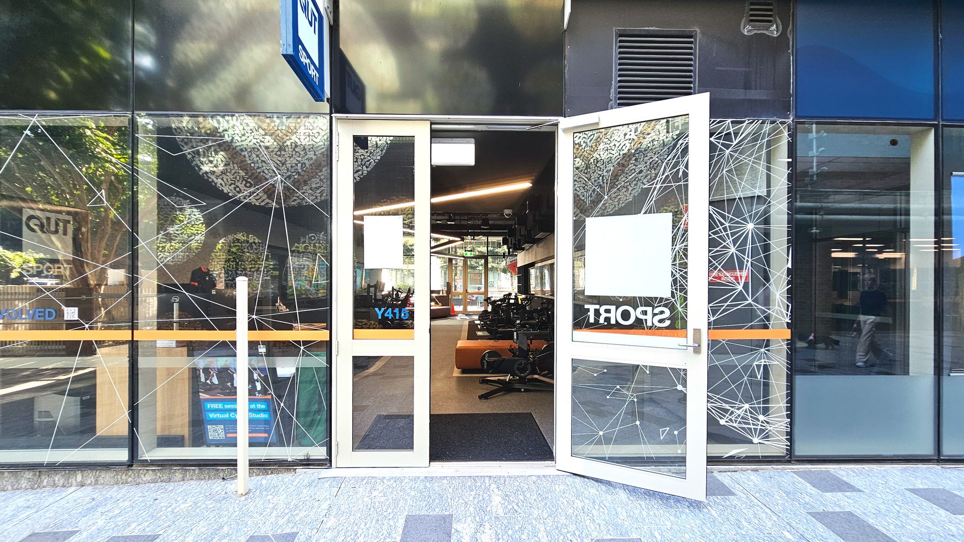 A modern entrance with glass doors open wide, revealing an interior with exercise equipment. The glass facade features geometric patterns and signage for 'QUT Sport'. The floor outside is tiled in grey, transitioning to a dark mat inside. A sign with 'Y415' is visible on the door. The space inside appears open with visible lighting fixtures and a reflective ceiling. A modern entrance with glass doors open wide, revealing an interior with exercise equipment. The glass facade features geometric patterns and signage for 'QUT Sport'. The floor outside is tiled in grey, transitioning to a dark mat inside. A sign with 'Y415' is visible on the door. The space inside appears open with visible lighting fixtures and a reflective ceiling.