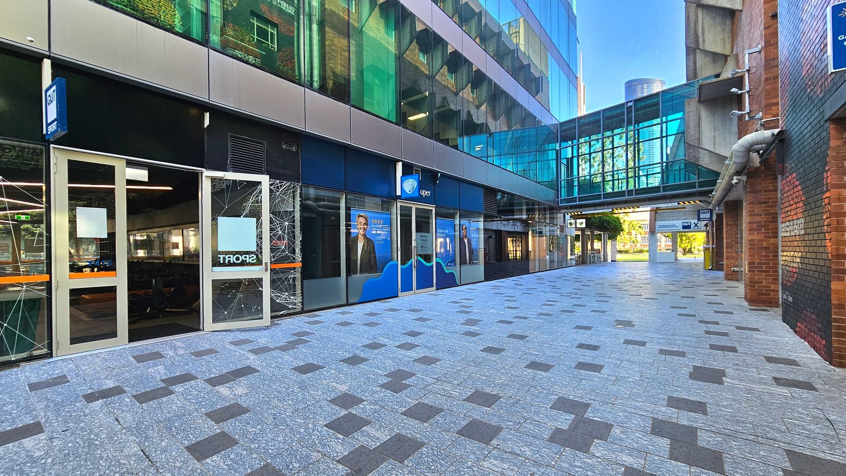 A modern urban walkway with a tiled floor featuring a pattern of grey and dark squares. On the left, a building with large glass windows displays signage for a sports facility. Above, a glass-enclosed pedestrian bridge connects two buildings. The right side has a brick wall with visible piping. In the distance, a green area is visible through the open end of the walkway. A modern urban walkway with a tiled floor featuring a pattern of grey and dark squares. On the left, a building with large glass windows displays signage for a sports facility. Above, a glass-enclosed pedestrian bridge connects two buildings. The right side has a brick wall with visible piping. In the distance, a green area is visible through the open end of the walkway.