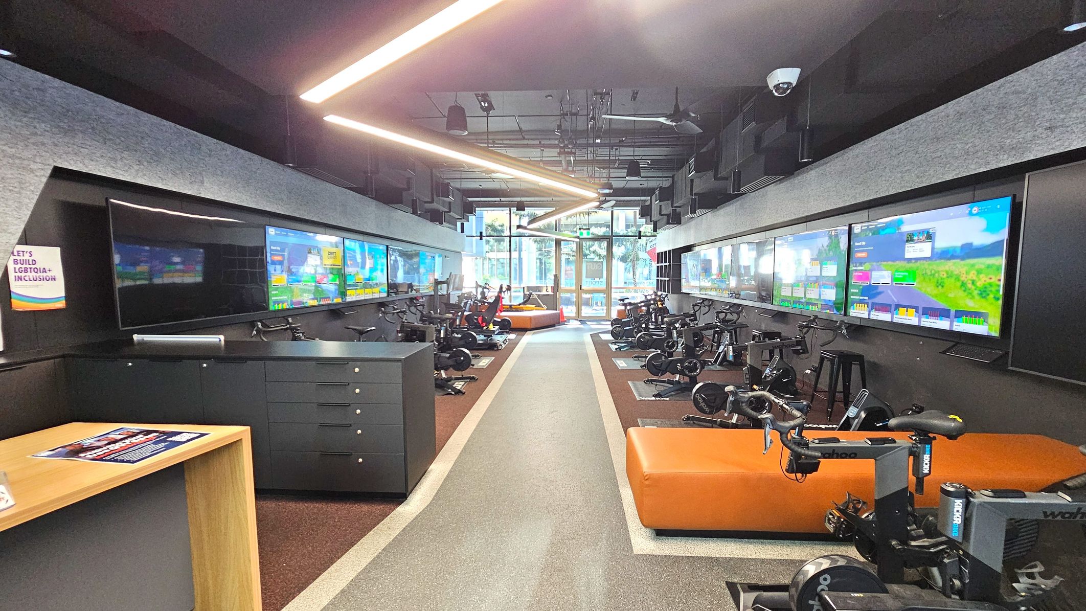 A modern indoor cycling studio with a central aisle flanked by stationary bikes on either side. Large screens displaying virtual cycling routes are mounted on the walls above the bikes. The floor is a mix of grey and brown materials, with a distinct central pathway. The ceiling features linear lighting and acoustic panels. At the far end, glass doors lead outside, and orange seating is visible near the entrance. A modern indoor cycling studio with a central aisle flanked by stationary bikes on either side. Large screens displaying virtual cycling routes are mounted on the walls above the bikes. The floor is a mix of grey and brown materials, with a distinct central pathway. The ceiling features linear lighting and acoustic panels. At the far end, glass doors lead outside, and orange seating is visible near the entrance.