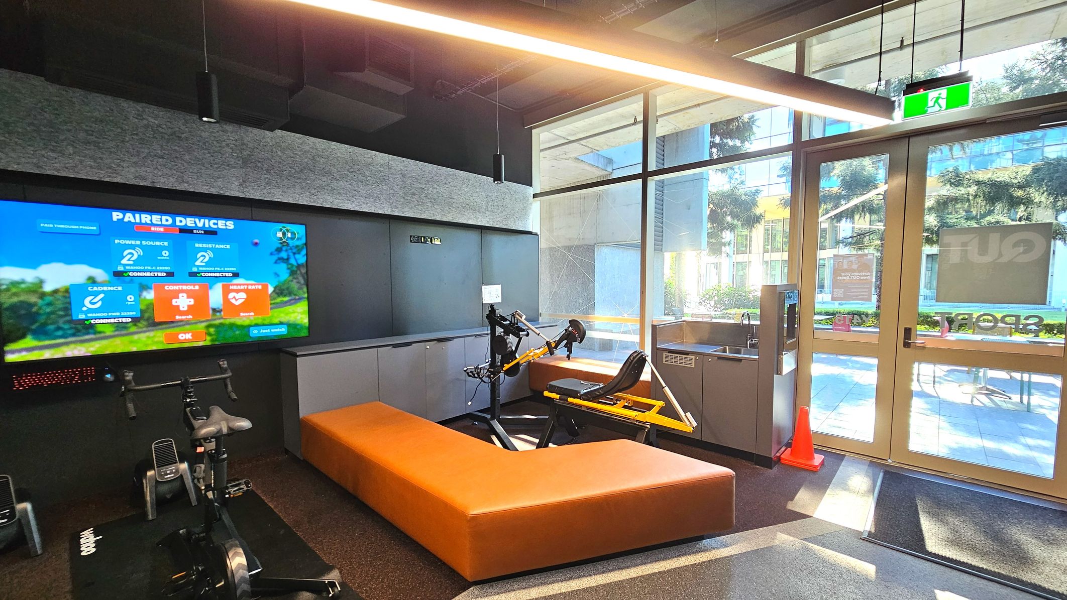 A modern indoor cycling area features a large screen displaying paired device information, mounted on a grey wall. Below, a stationary bike is positioned on a black mat. An L-shaped orange bench surrounds a yellow exercise machine. To the right, glass doors with an exit sign lead outside, flanked by a water dispenser and an orange cone. The floor transitions from dark carpet to a lighter mat near the entrance. A modern indoor cycling area features a large screen displaying paired device information, mounted on a grey wall. Below, a stationary bike is positioned on a black mat. An L-shaped orange bench surrounds a yellow exercise machine. To the right, glass doors with an exit sign lead outside, flanked by a water dispenser and an orange cone. The floor transitions from dark carpet to a lighter mat near the entrance.