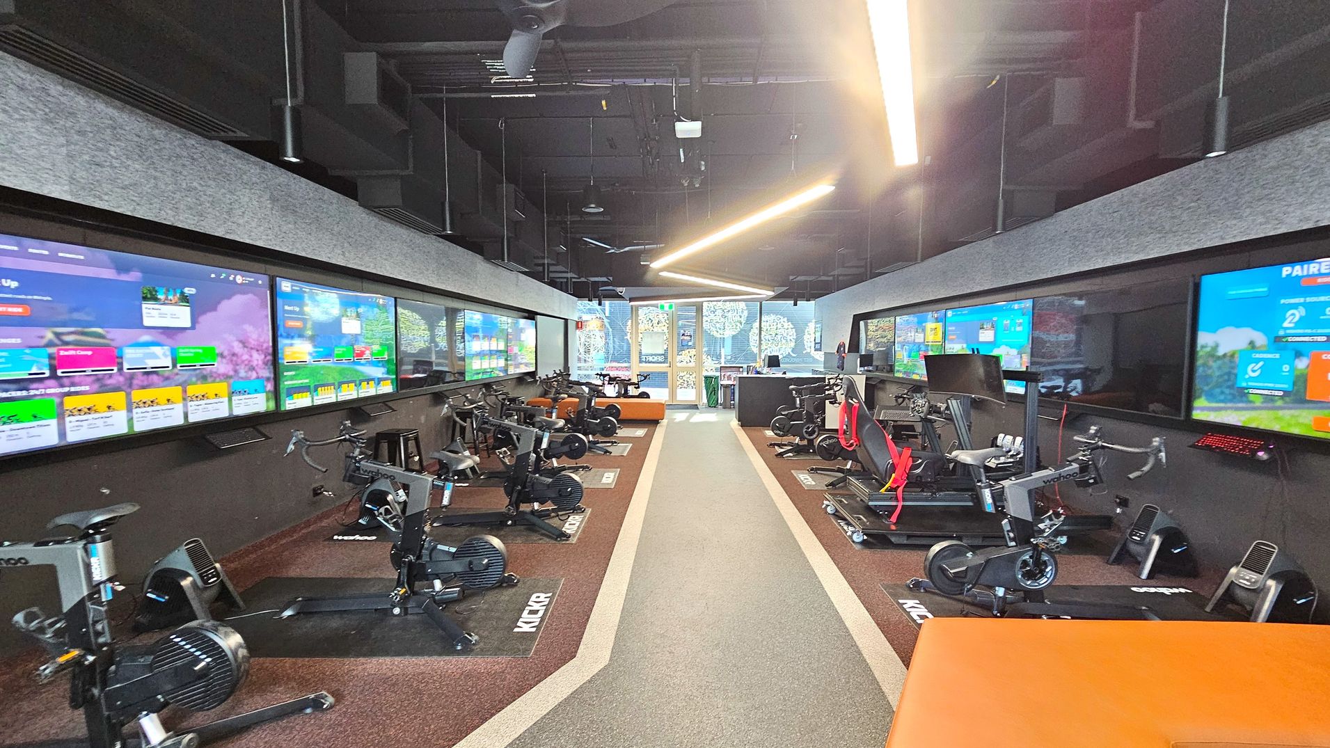 A modern indoor cycling studio with rows of stationary bikes on either side of a central aisle. Large digital screens line the walls above the bikes, displaying colourful graphics and information. The floor is carpeted with a central grey strip flanked by brown sections. The ceiling is high with exposed ductwork and linear lighting. At the far end, glass doors lead to another area, with decorative patterns visible on the glass. A modern indoor cycling studio with rows of stationary bikes on either side of a central aisle. Large digital screens line the walls above the bikes, displaying colourful graphics and information. The floor is carpeted with a central grey strip flanked by brown sections. The ceiling is high with exposed ductwork and linear lighting. At the far end, glass doors lead to another area, with decorative patterns visible on the glass.