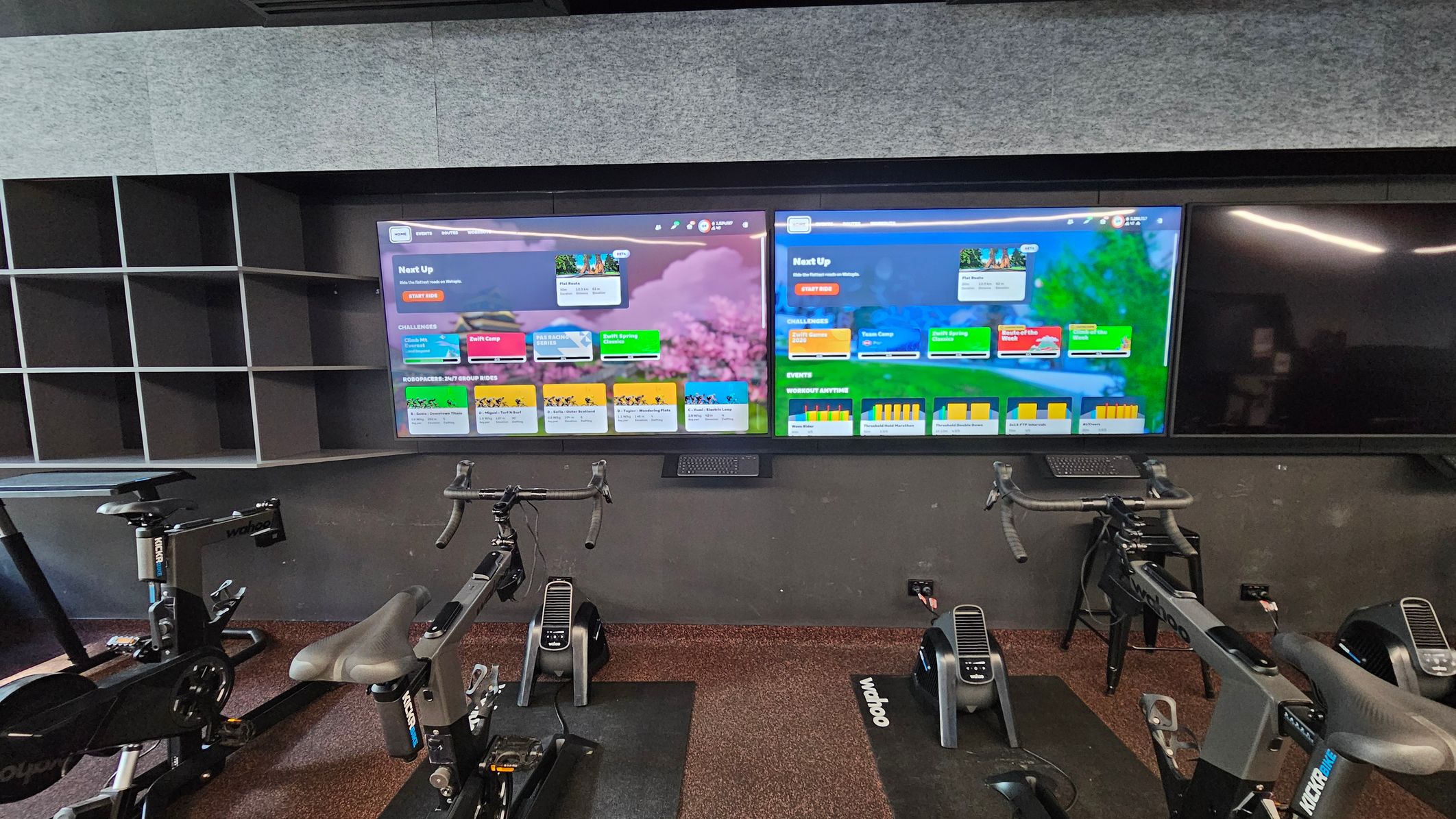 A modern indoor cycling studio featuring two large digital screens displaying colourful workout interfaces. Below the screens are three stationary bikes with sleek black frames and padded seats, positioned on dark mats. To the left, there is a wall-mounted shelving unit with multiple open compartments. The floor is covered in a speckled dark carpet, and the walls are lined with grey acoustic panels. A modern indoor cycling studio featuring two large digital screens displaying colourful workout interfaces. Below the screens are three stationary bikes with sleek black frames and padded seats, positioned on dark mats. To the left, there is a wall-mounted shelving unit with multiple open compartments. The floor is covered in a speckled dark carpet, and the walls are lined with grey acoustic panels.