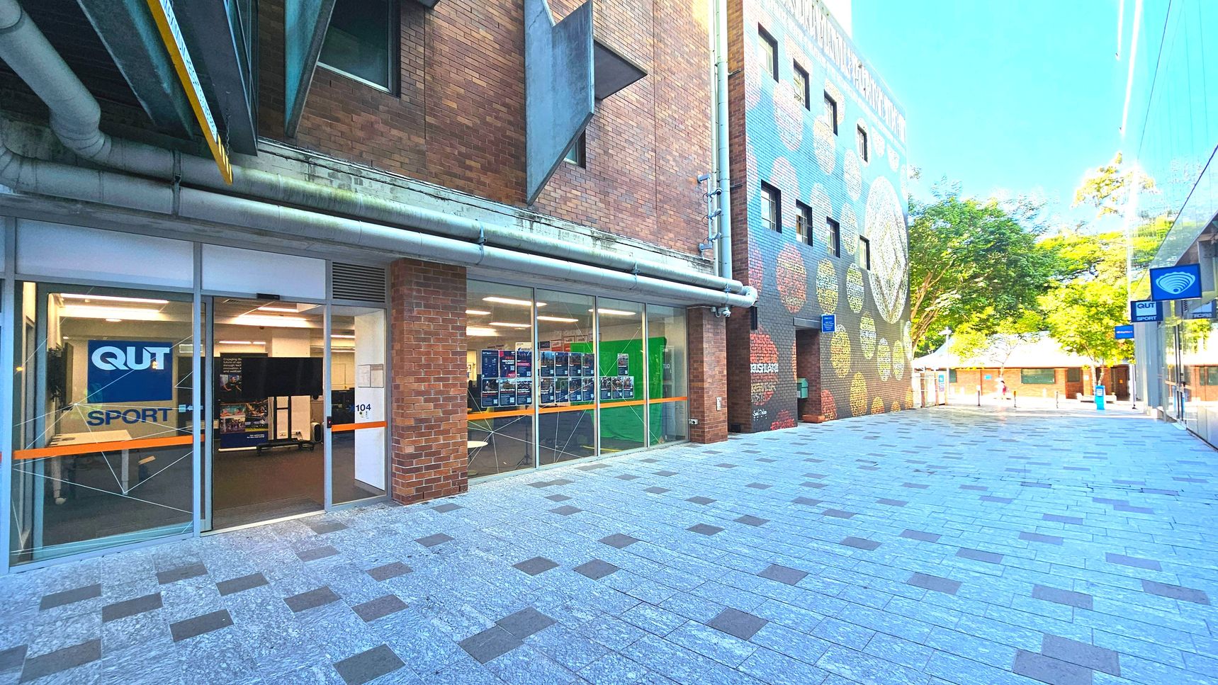 A brick building with large glass windows displays a sign reading 'QUT Sport'. The entrance is framed by a brick column. Adjacent is a colourful mural on another brick wall with circular patterns. The ground is paved with light grey tiles interspersed with darker squares. A blue sign with a Wi-Fi symbol hangs above. Trees and a small building are visible in the background, creating an open courtyard feel. A brick building with large glass windows displays a sign reading 'QUT Sport'. The entrance is framed by a brick column. Adjacent is a colourful mural on another brick wall with circular patterns. The ground is paved with light grey tiles interspersed with darker squares. A blue sign with a Wi-Fi symbol hangs above. Trees and a small building are visible in the background, creating an open courtyard feel.