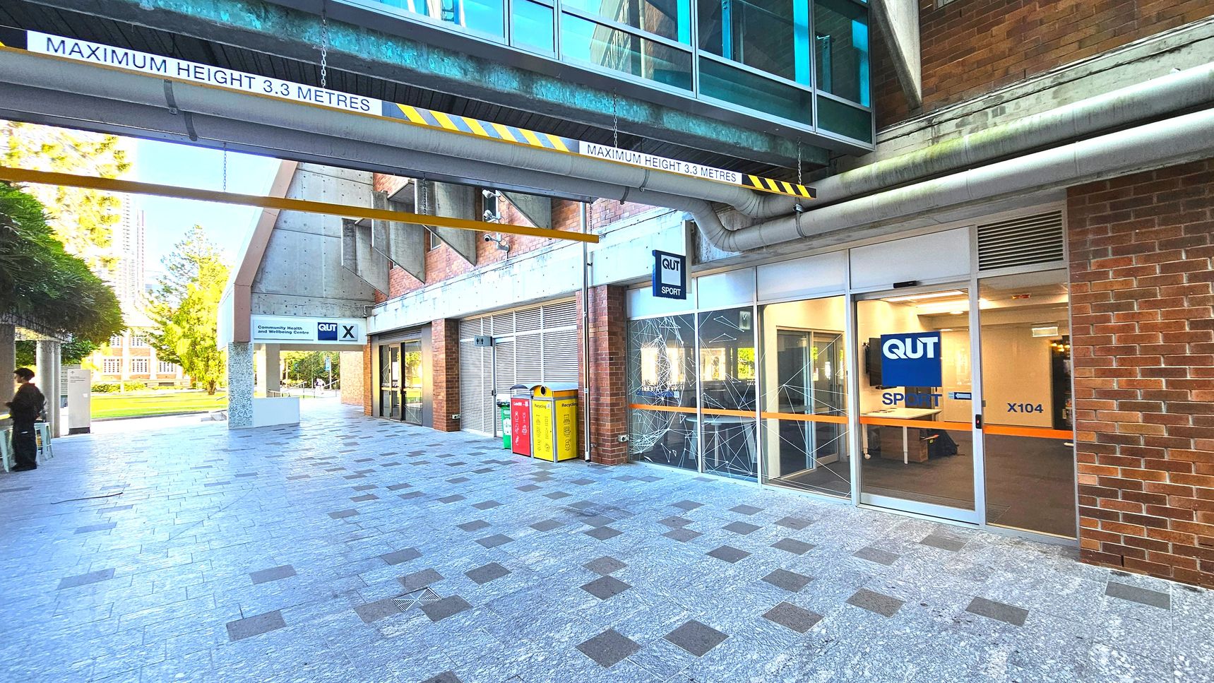 A covered walkway with a maximum height sign of 3.3 metres is visible, featuring a glass and metal overpass above. The ground is paved with grey and dark square tiles. On the right, a brick building with glass doors marked 'QUT Sport X104' is present. To the left, a sign for 'QUT' and bins in red, yellow, and green are seen. The area feels open with a view of a grassy area and trees in the background. A covered walkway with a maximum height sign of 3.3 metres is visible, featuring a glass and metal overpass above. The ground is paved with grey and dark square tiles. On the right, a brick building with glass doors marked 'QUT Sport X104' is present. To the left, a sign for 'QUT' and bins in red, yellow, and green are seen. The area feels open with a view of a grassy area and trees in the background.
