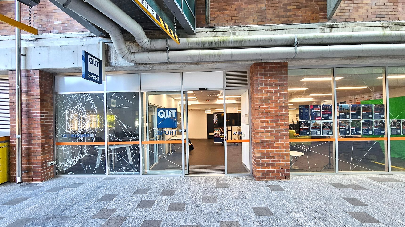 A brick building facade with large glass windows and a central entrance. The windows are decorated with white geometric patterns and an orange stripe. A sign reads 'QUT Sport' above the entrance. Inside, a well-lit room with posters on the walls is visible. The floor outside is a mix of grey and brown tiles. Overhead, large pipes run along the building, and a yellow clearance sign hangs above. A brick building facade with large glass windows and a central entrance. The windows are decorated with white geometric patterns and an orange stripe. A sign reads 'QUT Sport' above the entrance. Inside, a well-lit room with posters on the walls is visible. The floor outside is a mix of grey and brown tiles. Overhead, large pipes run along the building, and a yellow clearance sign hangs above.