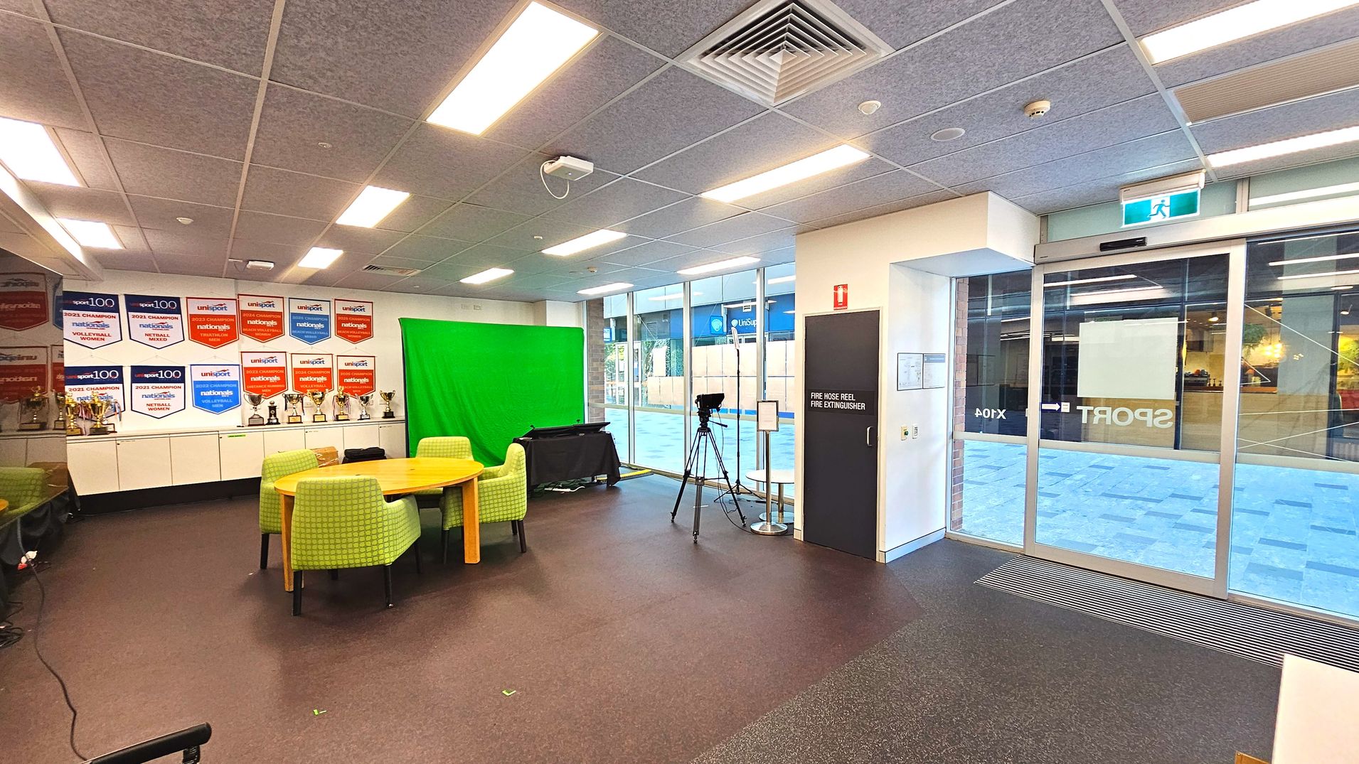 The room features a wall with numerous sports banners and trophies on white cabinets. A green screen is set up beside a round wooden table surrounded by green upholstered chairs. A camera on a tripod is positioned near a glass sliding door, which has a visible exit sign above. The floor is carpeted, and the ceiling has recessed lighting and ventilation panels. A door marked 'Fire Hose Reel Fire Extinguisher' is on the right. The room features a wall with numerous sports banners and trophies on white cabinets. A green screen is set up beside a round wooden table surrounded by green upholstered chairs. A camera on a tripod is positioned near a glass sliding door, which has a visible exit sign above. The floor is carpeted, and the ceiling has recessed lighting and ventilation panels. A door marked 'Fire Hose Reel Fire Extinguisher' is on the right.