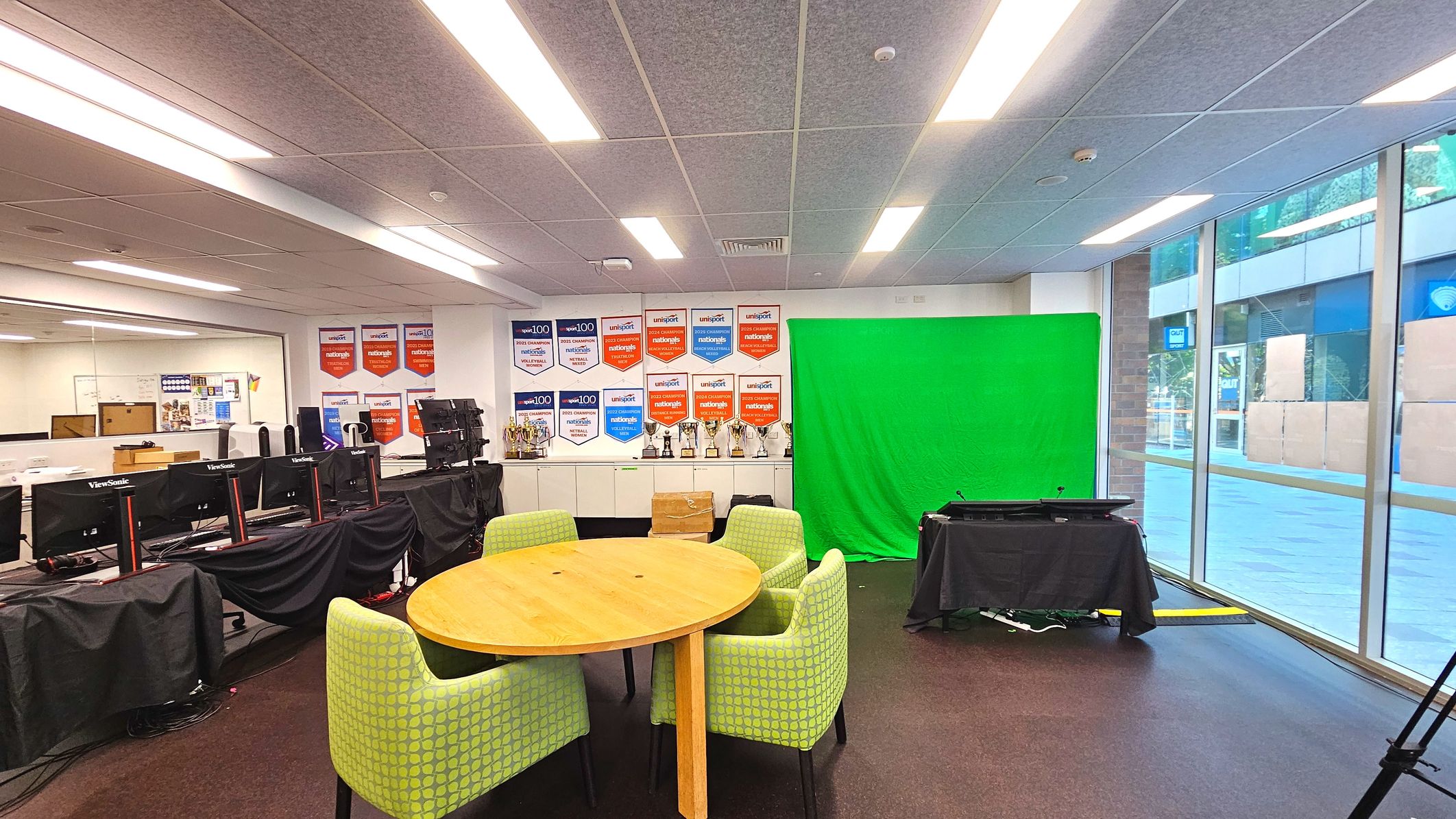 An indoor space featuring a round wooden table surrounded by four green upholstered chairs with a geometric pattern. To the right, a large green screen is set up against a wall adorned with numerous award banners and trophies on a shelf. Several computer monitors are covered with black cloths on the left. The room has a low ceiling with fluorescent lighting and large glass windows on the right, offering a view of an outdoor area. An indoor space featuring a round wooden table surrounded by four green upholstered chairs with a geometric pattern. To the right, a large green screen is set up against a wall adorned with numerous award banners and trophies on a shelf. Several computer monitors are covered with black cloths on the left. The room has a low ceiling with fluorescent lighting and large glass windows on the right, offering a view of an outdoor area.