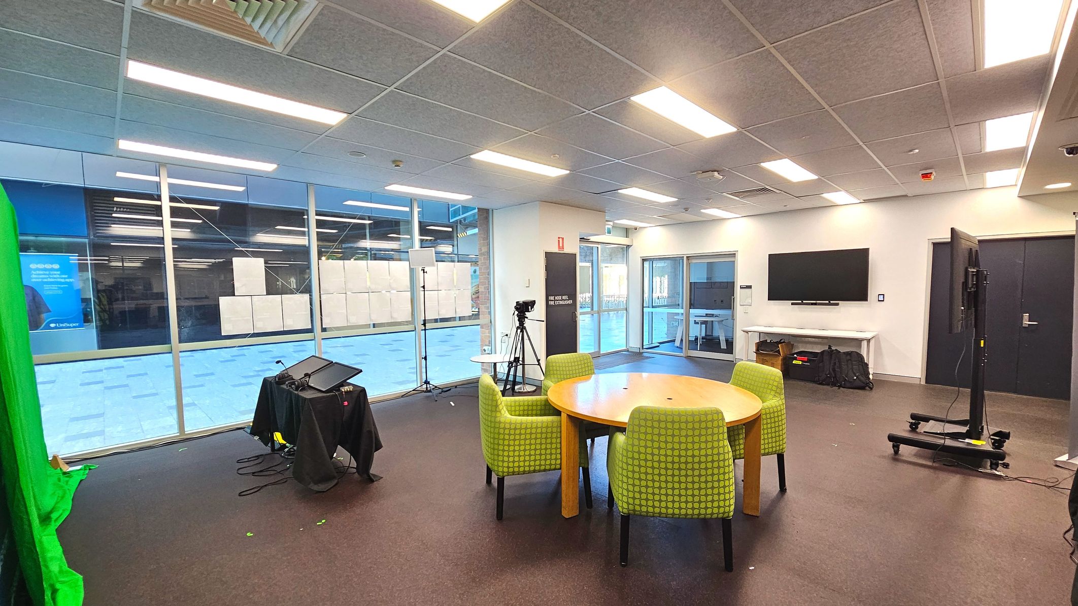 A modern room with a large glass wall on the left, featuring a green screen setup and a camera on a tripod. In the centre, a round wooden table is surrounded by four green upholstered chairs. A large flat-screen TV is mounted on the right wall above a white console table. The floor is carpeted in dark grey, and the ceiling has recessed lighting. A set of double glass doors is visible in the background. A modern room with a large glass wall on the left, featuring a green screen setup and a camera on a tripod. In the centre, a round wooden table is surrounded by four green upholstered chairs. A large flat-screen TV is mounted on the right wall above a white console table. The floor is carpeted in dark grey, and the ceiling has recessed lighting. A set of double glass doors is visible in the background.