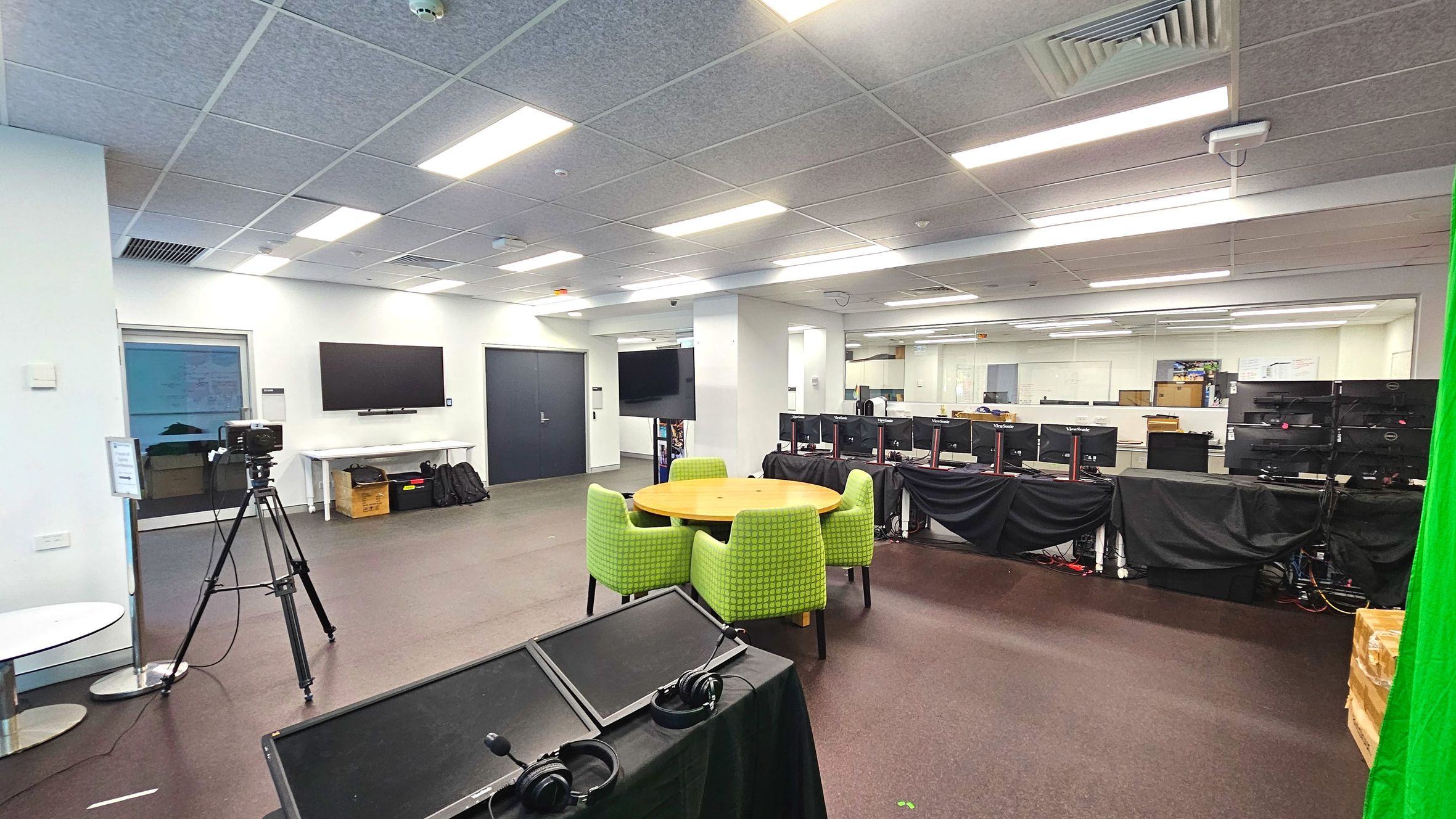 A spacious room with a modern office layout. In the foreground, a round wooden table is surrounded by four green upholstered chairs. To the left, a camera on a tripod faces a wall-mounted screen above a white table with bags underneath. Several computer monitors are lined up on desks covered with black cloths. The ceiling features recessed lighting and air vents. The floor is carpeted, and a green screen is partially visible on the right. A spacious room with a modern office layout. In the foreground, a round wooden table is surrounded by four green upholstered chairs. To the left, a camera on a tripod faces a wall-mounted screen above a white table with bags underneath. Several computer monitors are lined up on desks covered with black cloths. The ceiling features recessed lighting and air vents. The floor is carpeted, and a green screen is partially visible on the right.
