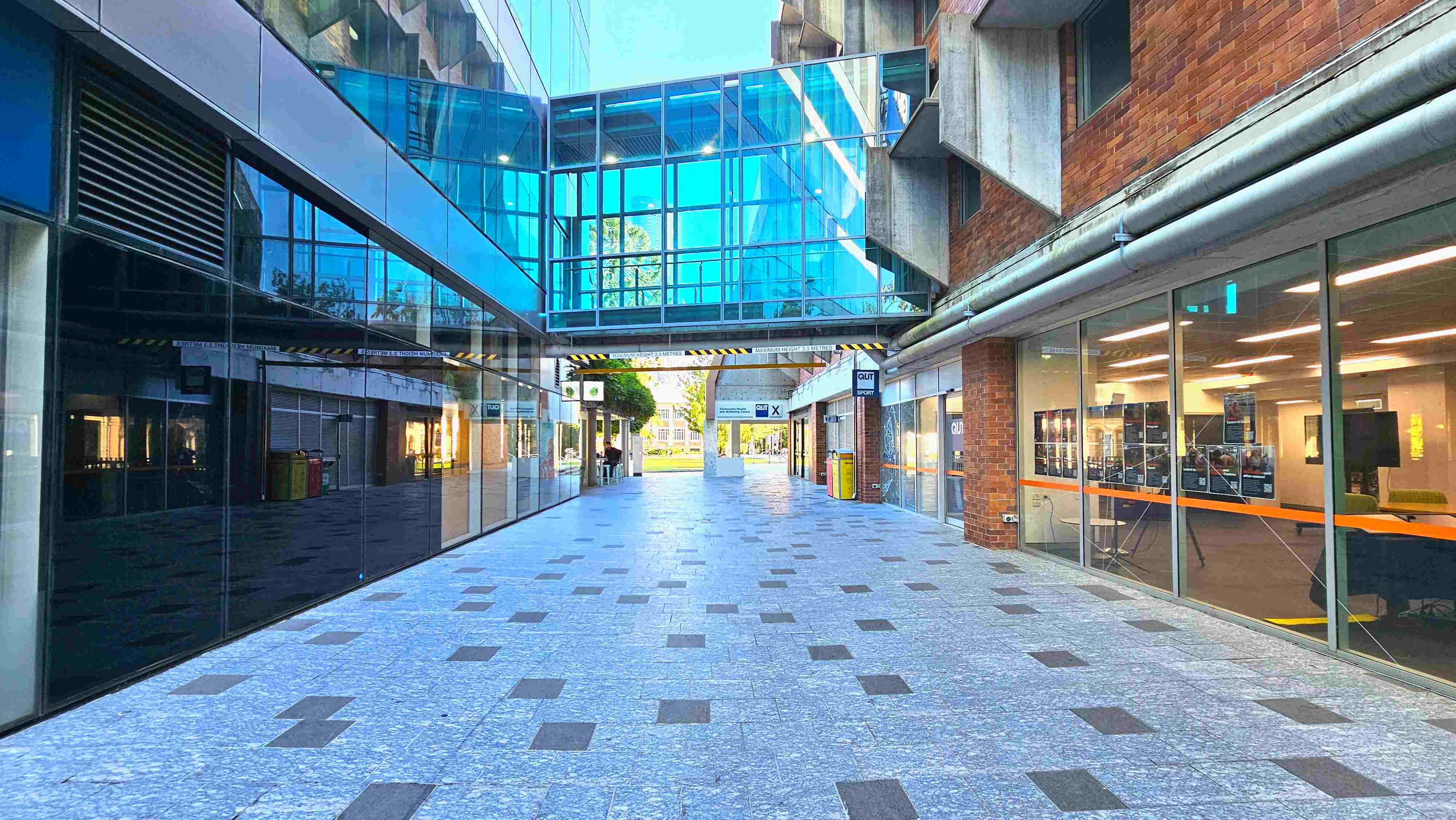 A modern walkway with a polished stone floor featuring square patterns, flanked by glass-fronted buildings. On the left, reflective glass panels mirror the scene, while the right side has brick walls and large windows displaying interior spaces. A blue-tinted glass skybridge connects the buildings overhead. Signs and bins are visible along the path, which leads to an open area with greenery in the distance. A modern walkway with a polished stone floor featuring square patterns, flanked by glass-fronted buildings. On the left, reflective glass panels mirror the scene, while the right side has brick walls and large windows displaying interior spaces. A blue-tinted glass skybridge connects the buildings overhead. Signs and bins are visible along the path, which leads to an open area with greenery in the distance.