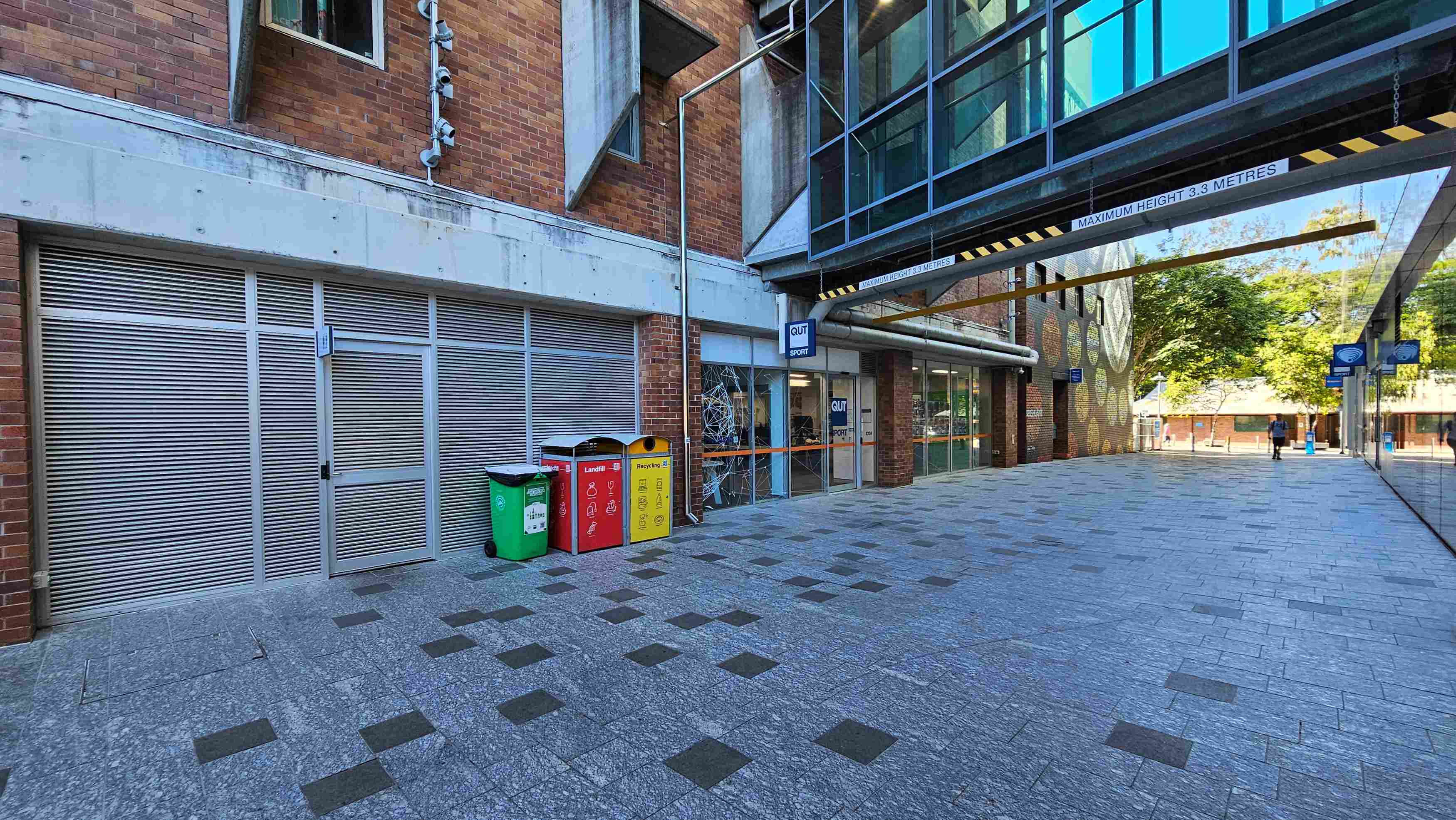 A modern urban walkway with a brick building on the left featuring metal louvred doors and a glass entrance labelled 'QUT Sport'. Three colourful bins for waste segregation are positioned nearby. Overhead, a glass bridge connects buildings, marked with a 'Maximum Height 3.3 Metres' sign. The ground is paved with grey tiles interspersed with darker squares. The area feels open, with trees visible in the background. A modern urban walkway with a brick building on the left featuring metal louvred doors and a glass entrance labelled 'QUT Sport'. Three colourful bins for waste segregation are positioned nearby. Overhead, a glass bridge connects buildings, marked with a 'Maximum Height 3.3 Metres' sign. The ground is paved with grey tiles interspersed with darker squares. The area feels open, with trees visible in the background.